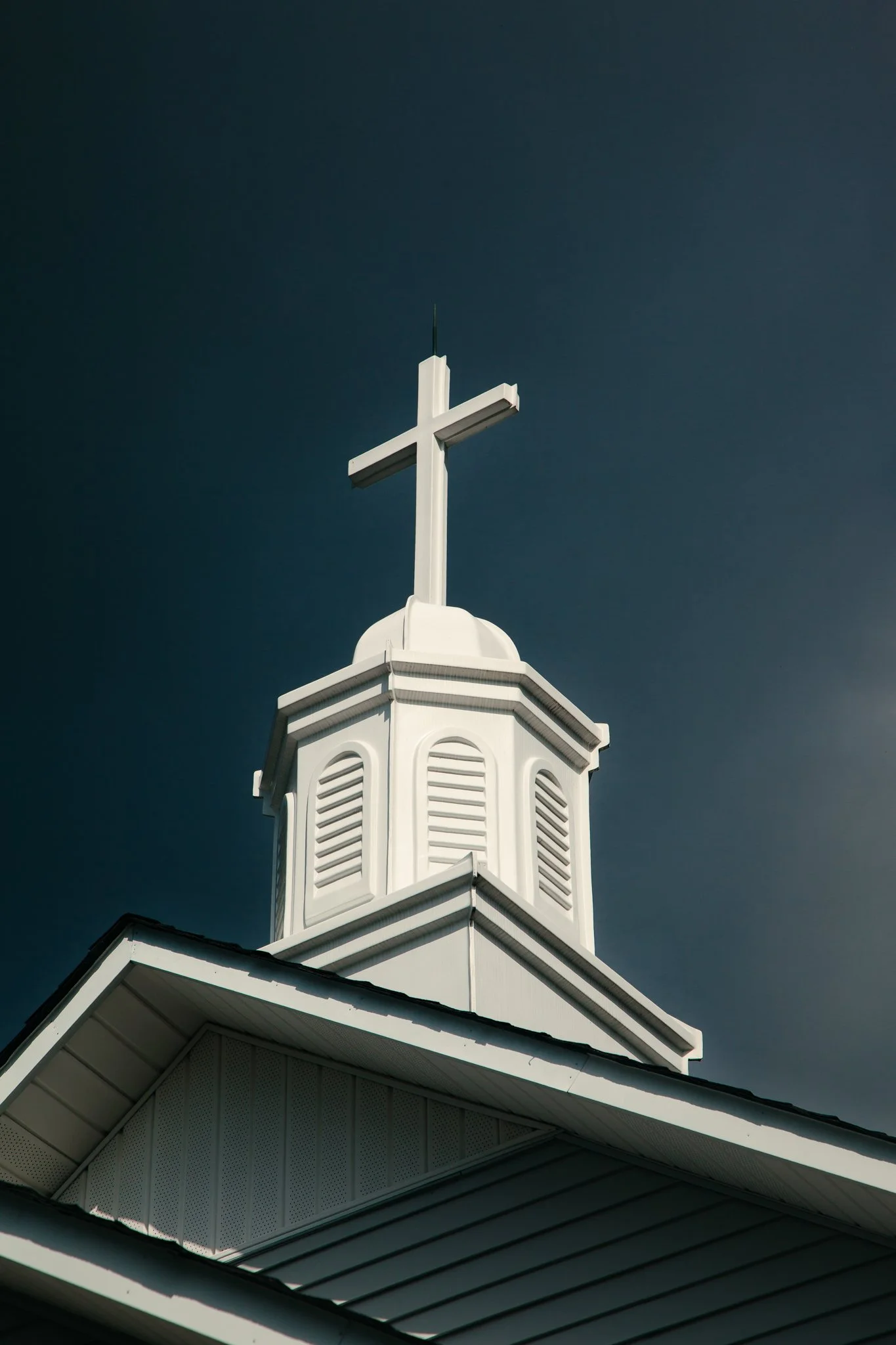 Close-up of a white church steeple with a cross on top, against a dark blue sky.