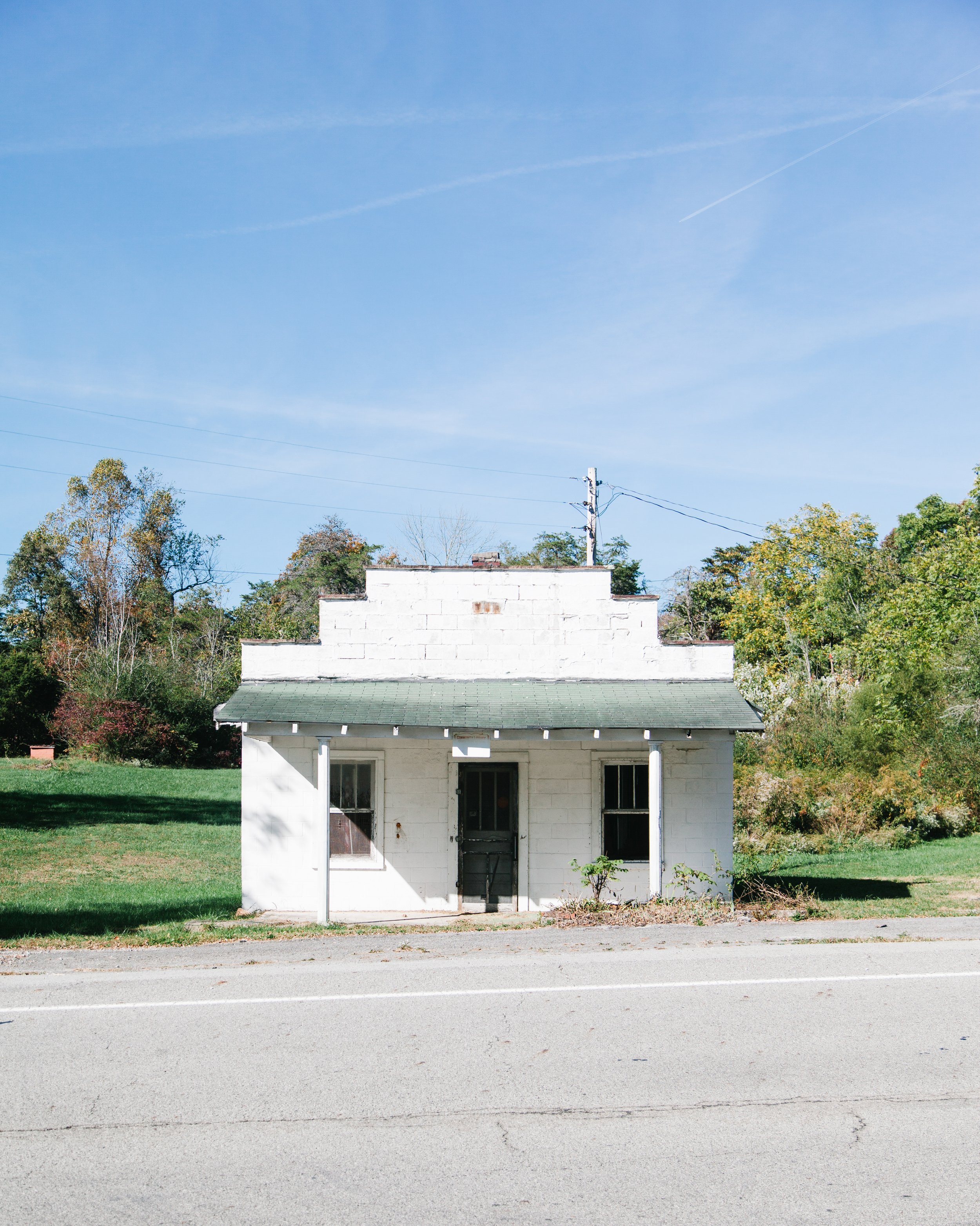 Small white house with a porch, black door, and windows, situated on a grassy area with asphalt road in the foreground, surrounded by trees with green and autumn-colored leaves, under a clear blue sky.