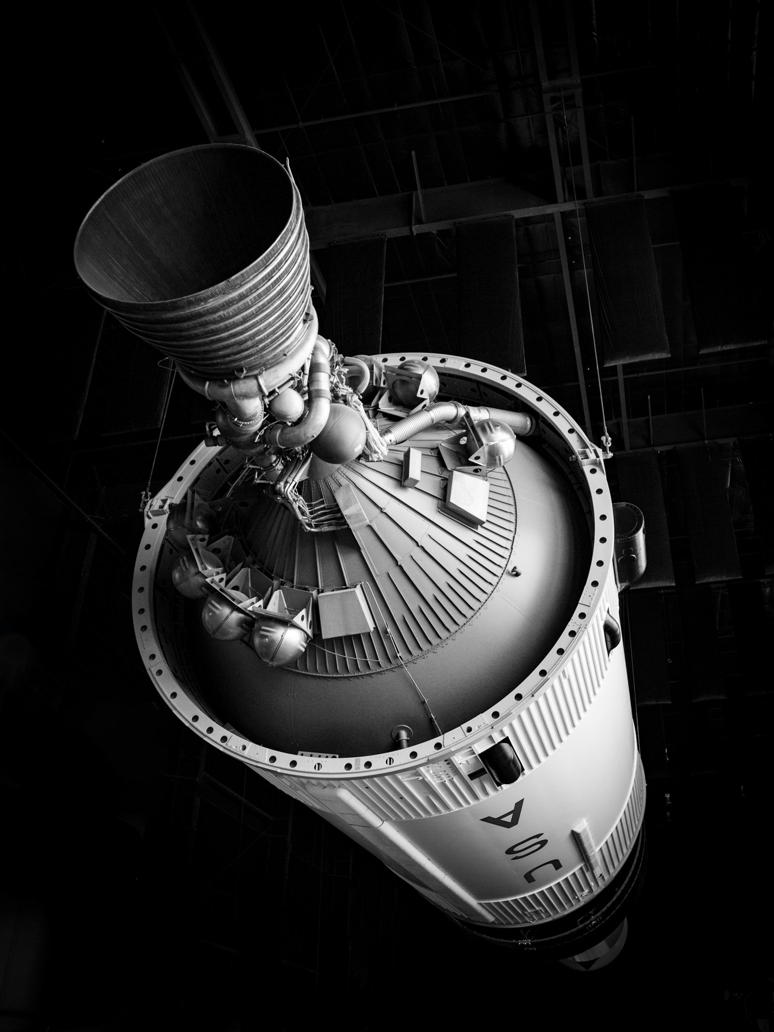A close-up view of a large rocket engine or thruster with intricate piping and mechanical parts, hanging vertically in a dark industrial setting.