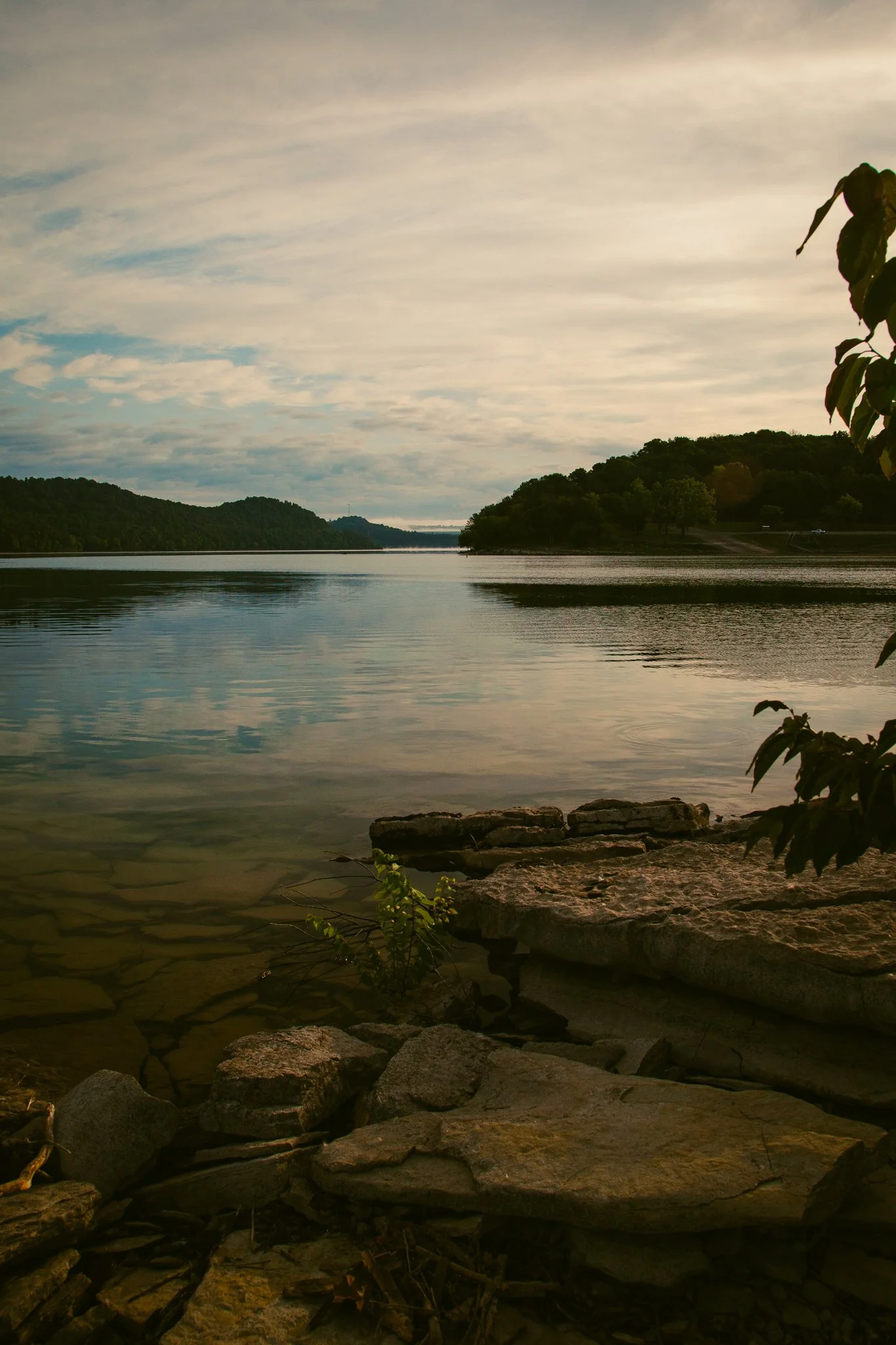 View of a calm river with surrounding green trees and hills, under a partly cloudy sky with soft sunlight.