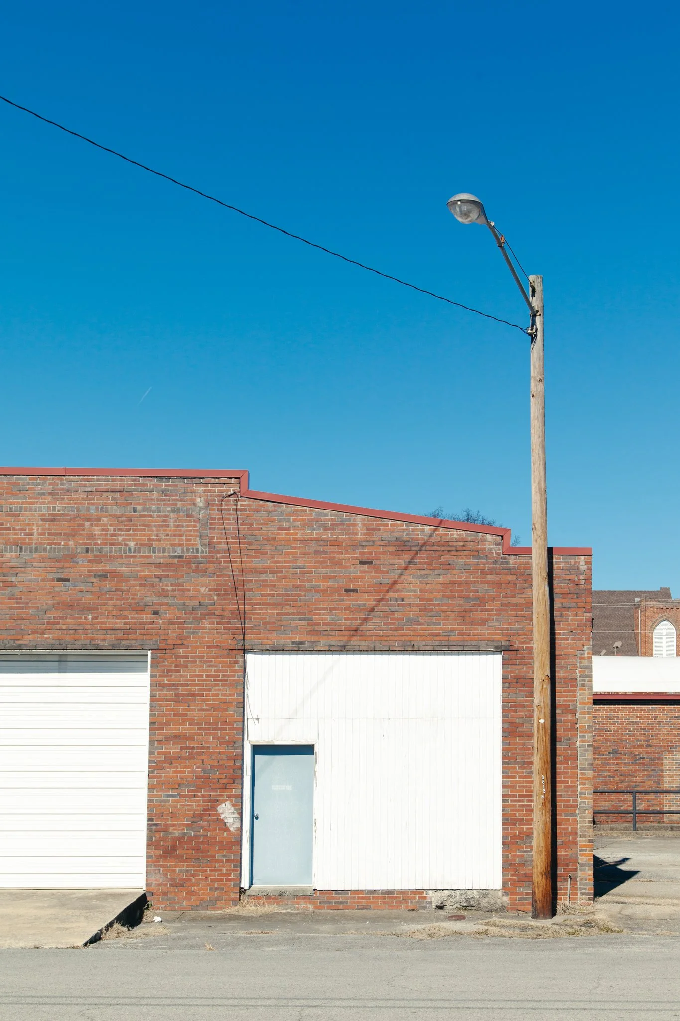A brick building with closed garage doors, a blue door with a window, a tall wooden utility pole, and a streetlamp against a clear blue sky.