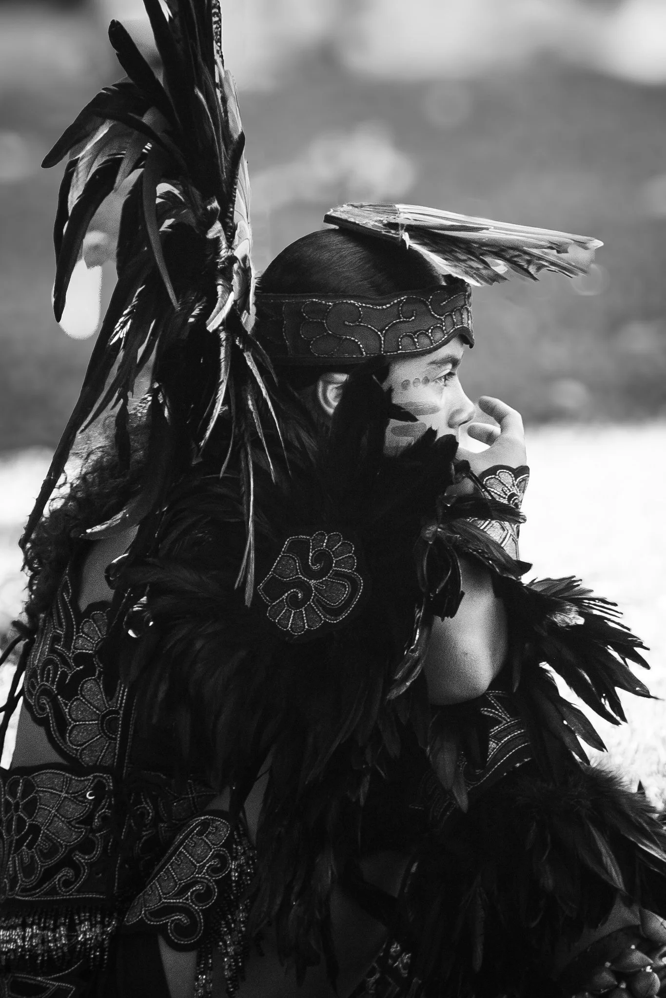 A person dressed in traditional indigenous attire, wearing a headpiece decorated with long feathers and an embroidered band, with face paint and detailed beadwork on their clothing.