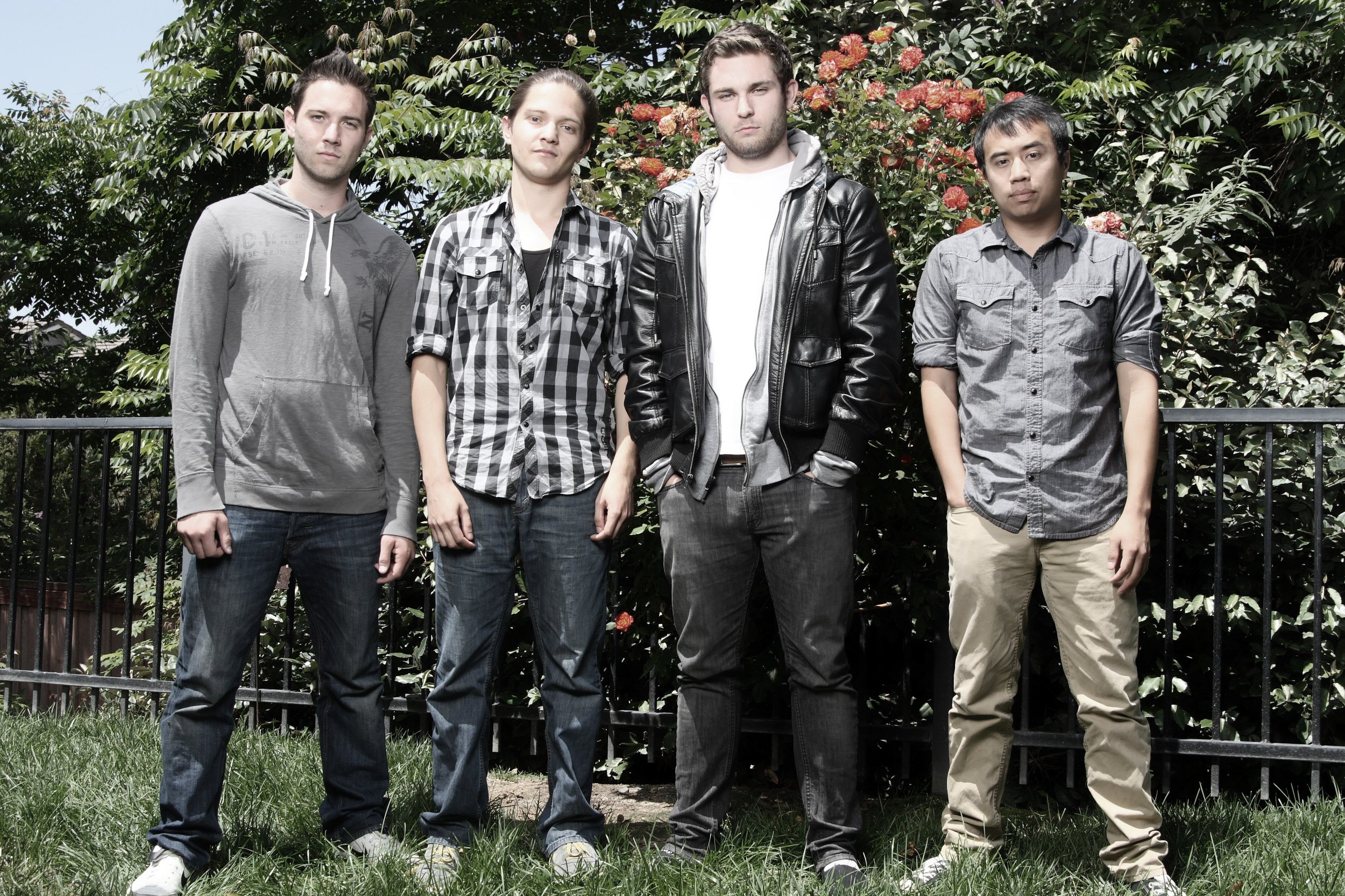 A group of five young men standing outdoors in front of green foliage, wearing casual clothing and looking at the camera.
