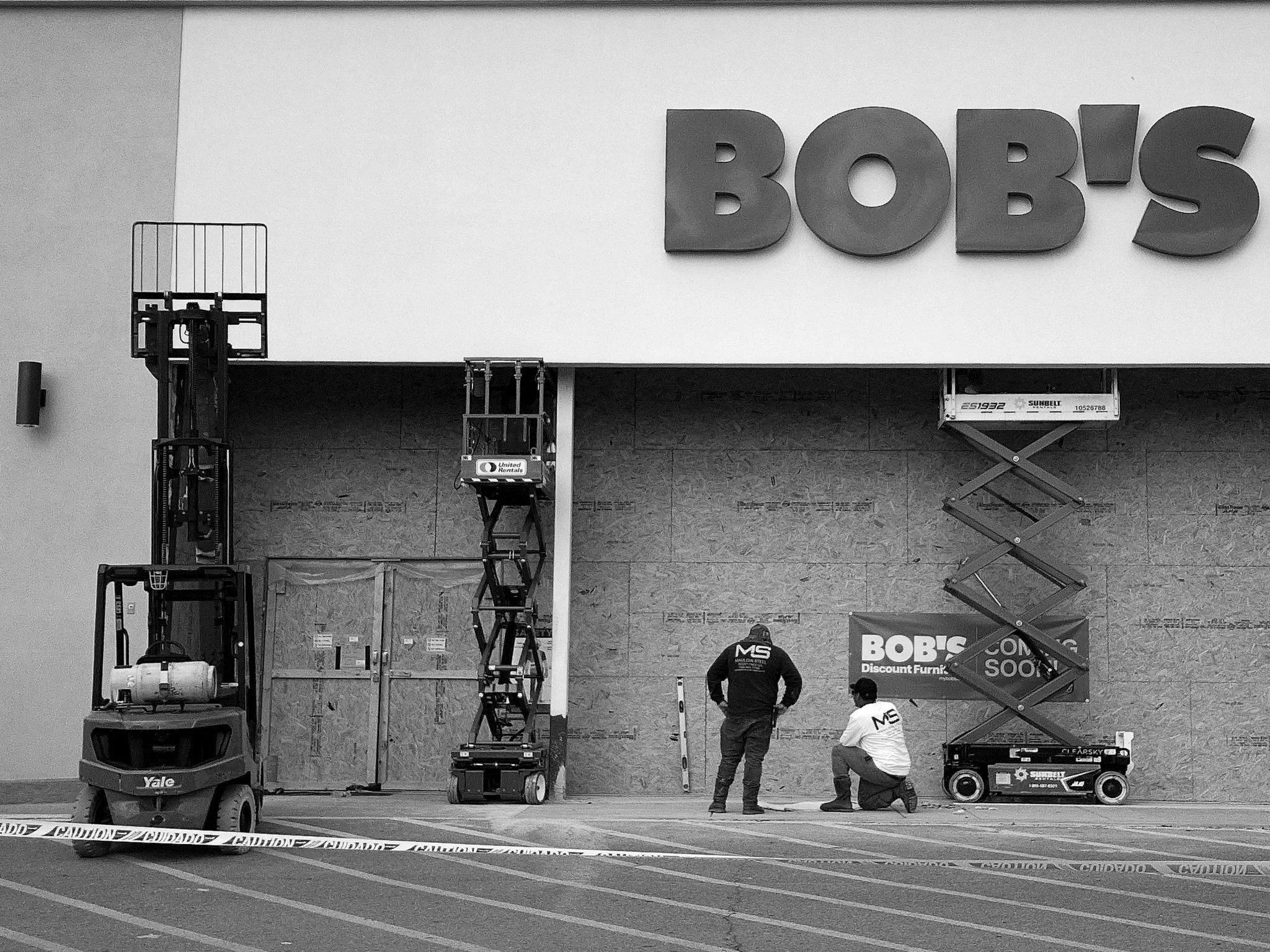 A store under construction or renovation with workers and equipment. Large letters spelling 'BOB'S' are mounted on the wall, and construction lifts and scaffolding are present in the scene.