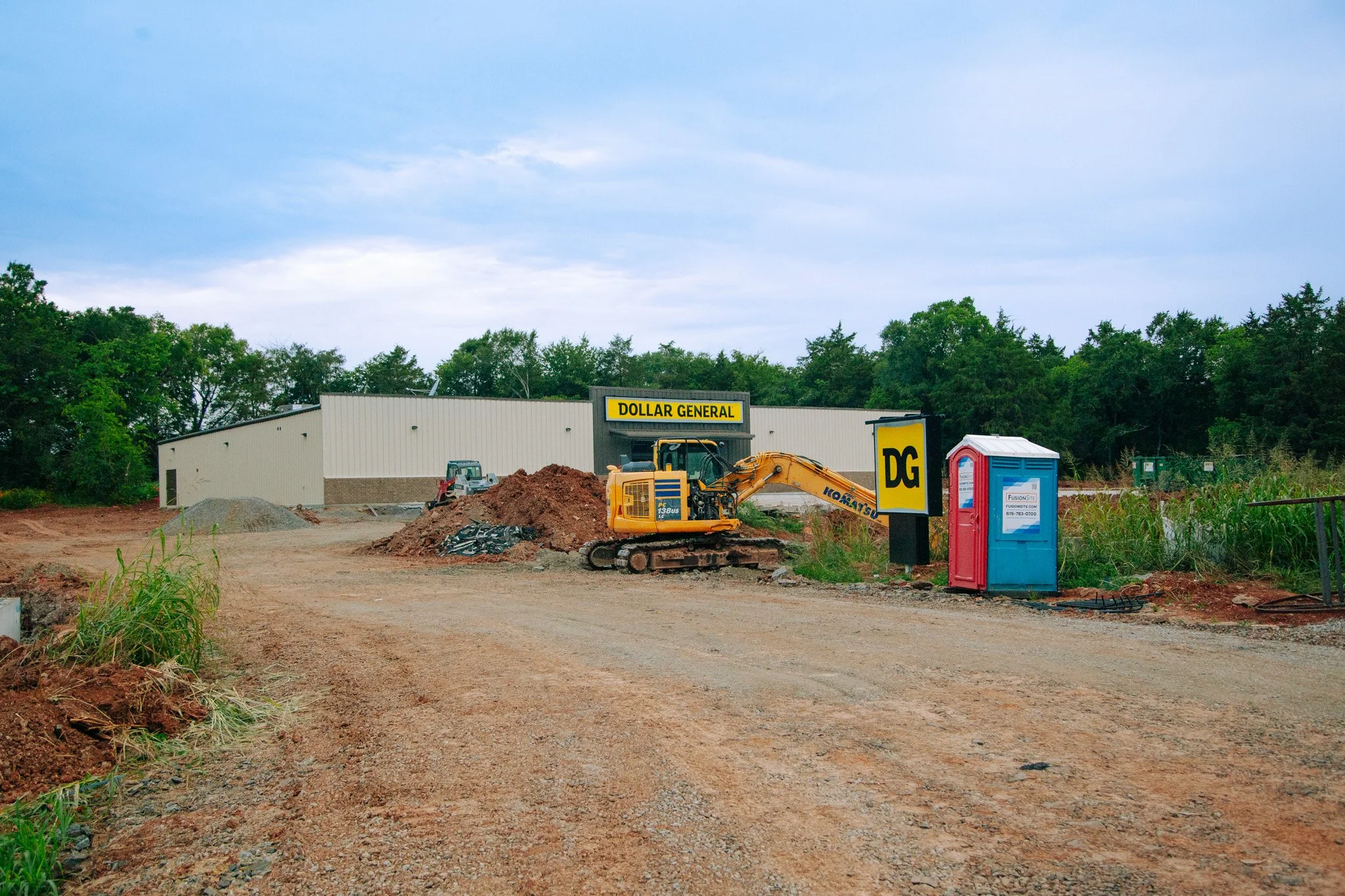 Construction site with an excavator, a portable toilet, and a Dollar General store in the background, surrounded by trees and a dirt ground.