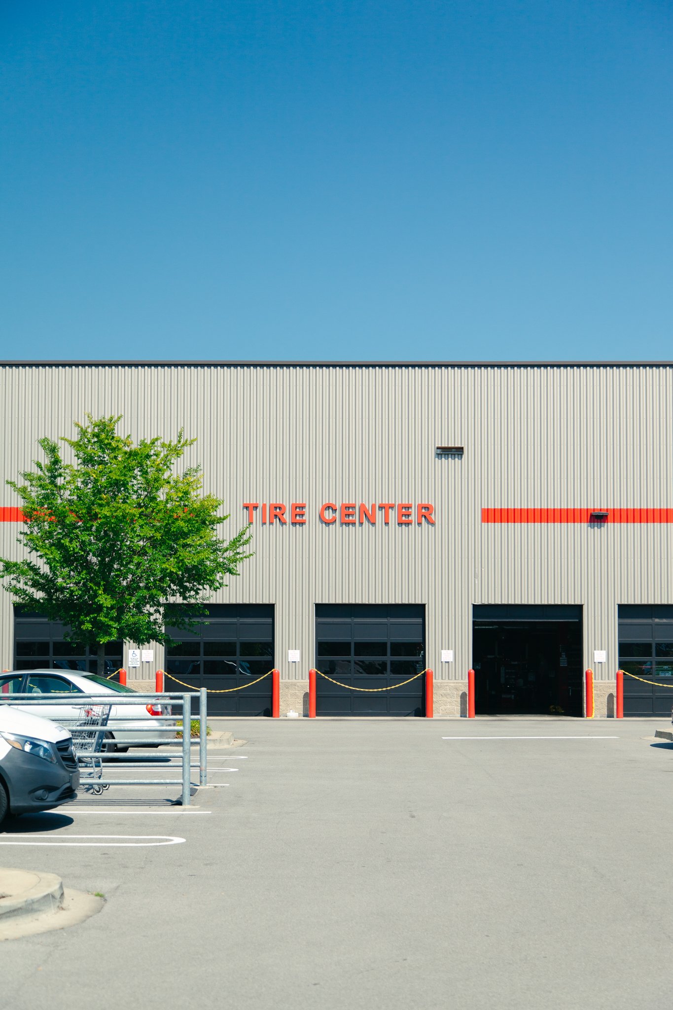 Exterior view of a large tire store called Tire Center with a gray metal facade, red signage, and a parking lot with cars, trees, and orange traffic cones.