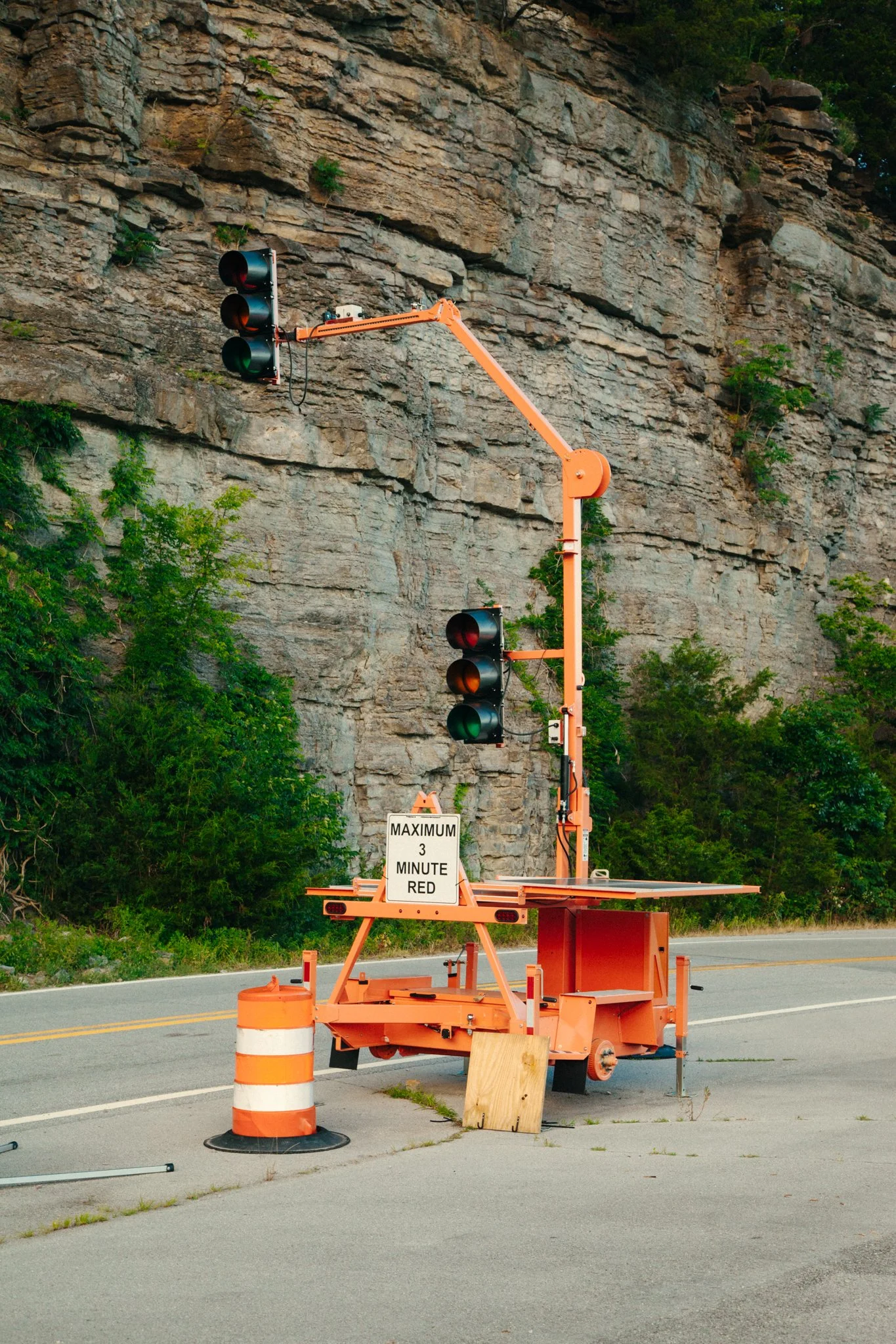 A portable traffic signal with red lights activated, mounted on an orange trailer near a rocky hillside. A sign on the trailer reads "Maximum 3 Minute Red." Orange and white traffic cones are surrounding the setup. There are trees and a two-lane road