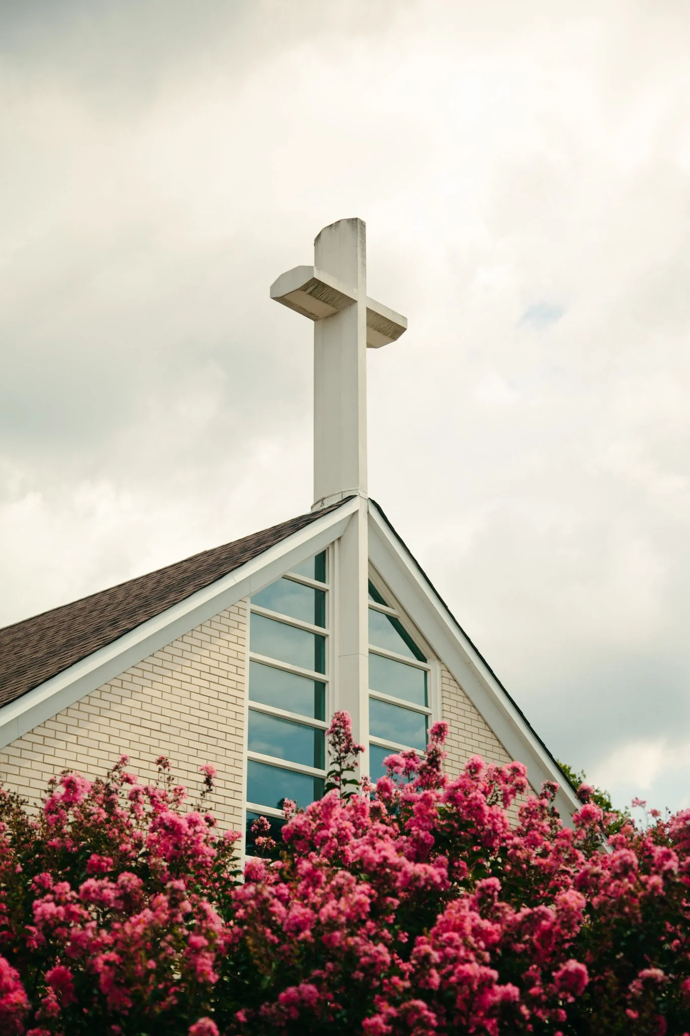 A church with a white cross on its roof, brown shingle roof, and large windows, surrounded by pink blooming flowers and a cloudy sky.