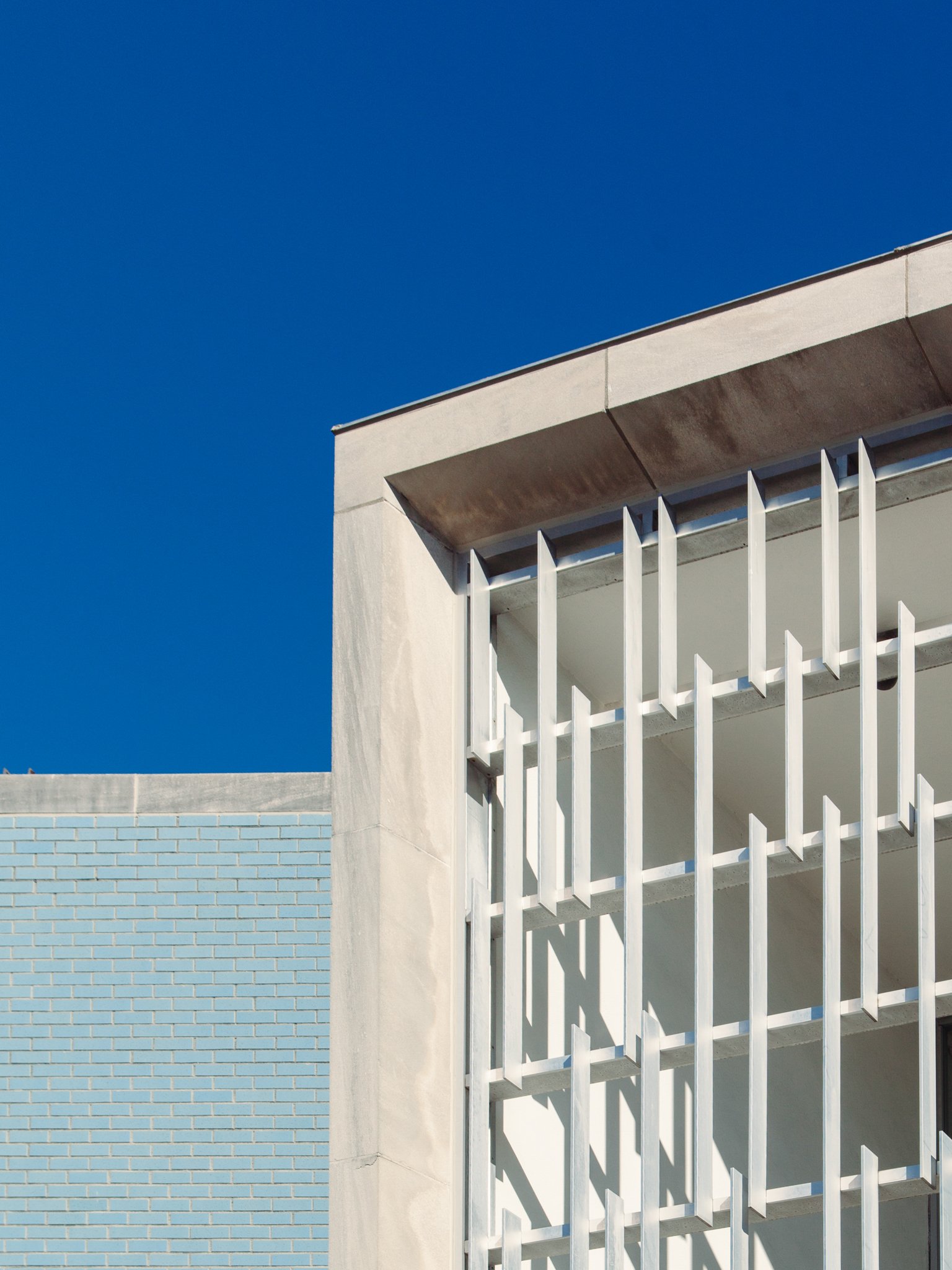 Part of a modern building with a white exterior, vertical white slats on the balcony, a light blue brick wall, and a clear blue sky.
