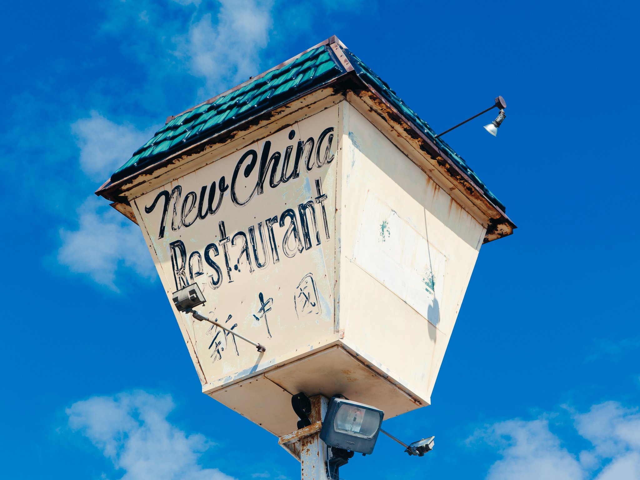 Close-up of a weathered, white, square sign with black cursive writing that reads 'Hearld Restaurant', attached to a rusty pole, under a bright blue sky with a few clouds. A small, adjustable floodlight is mounted on the top right corner of the sign.