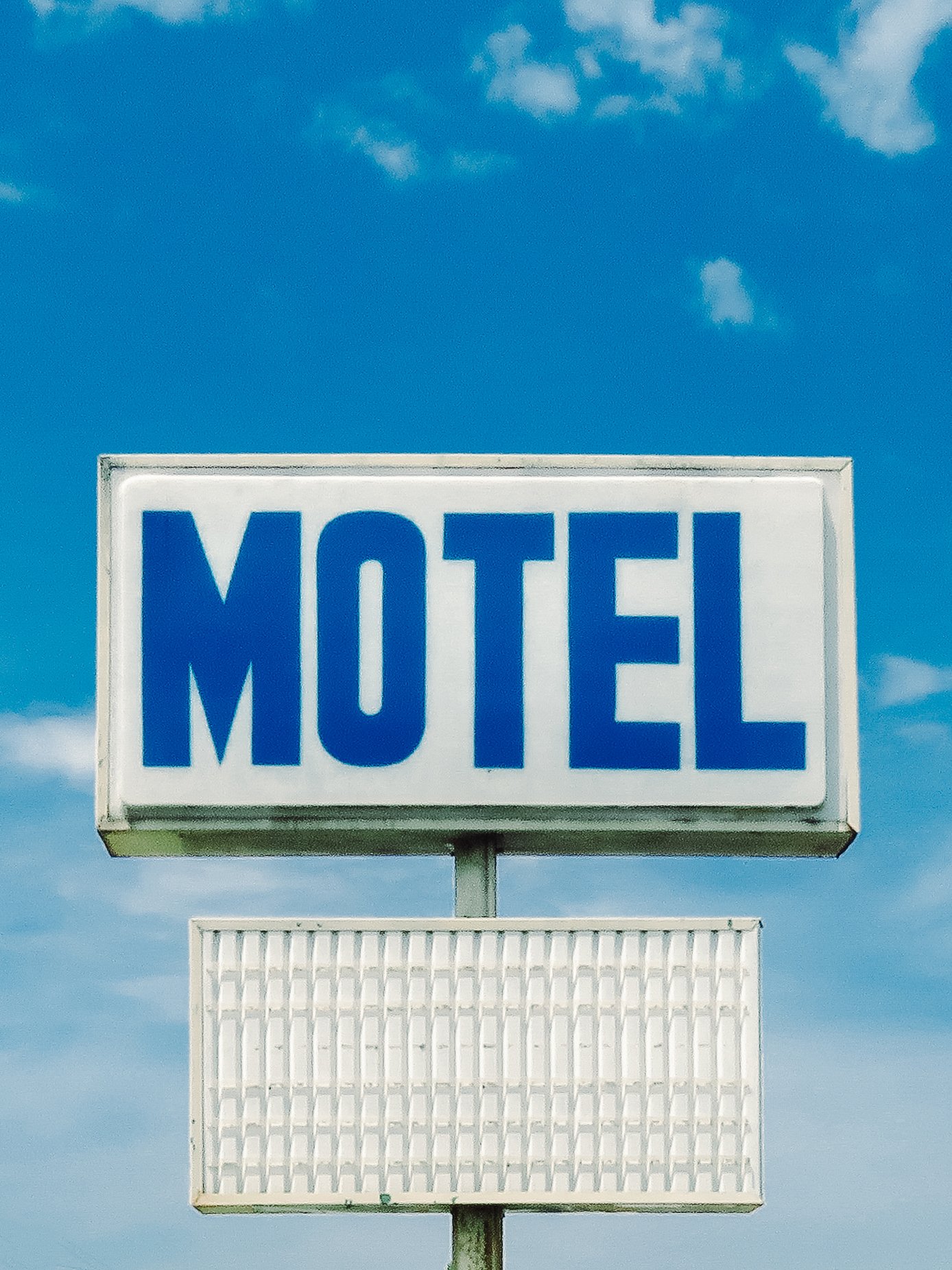 A bright blue sky with scattered white clouds and a large sign that says 'MOTEL' in blue uppercase letters on a white background, mounted on a pole.