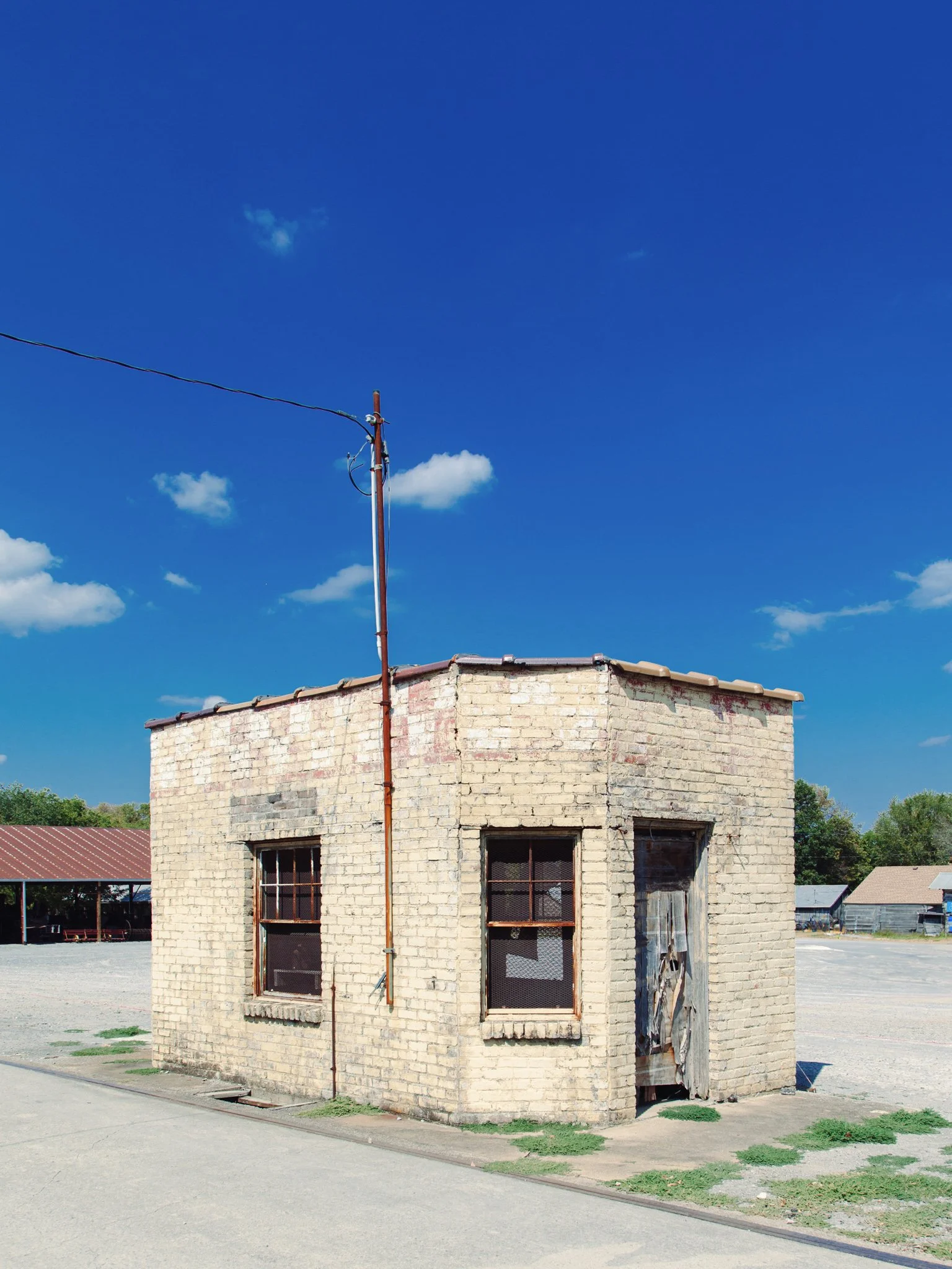 A small, abandoned brick building with boarded-up windows and peeling paint, situated on a paved lot under a bright blue sky with a few clouds. A utility pole with wires is next to the building.