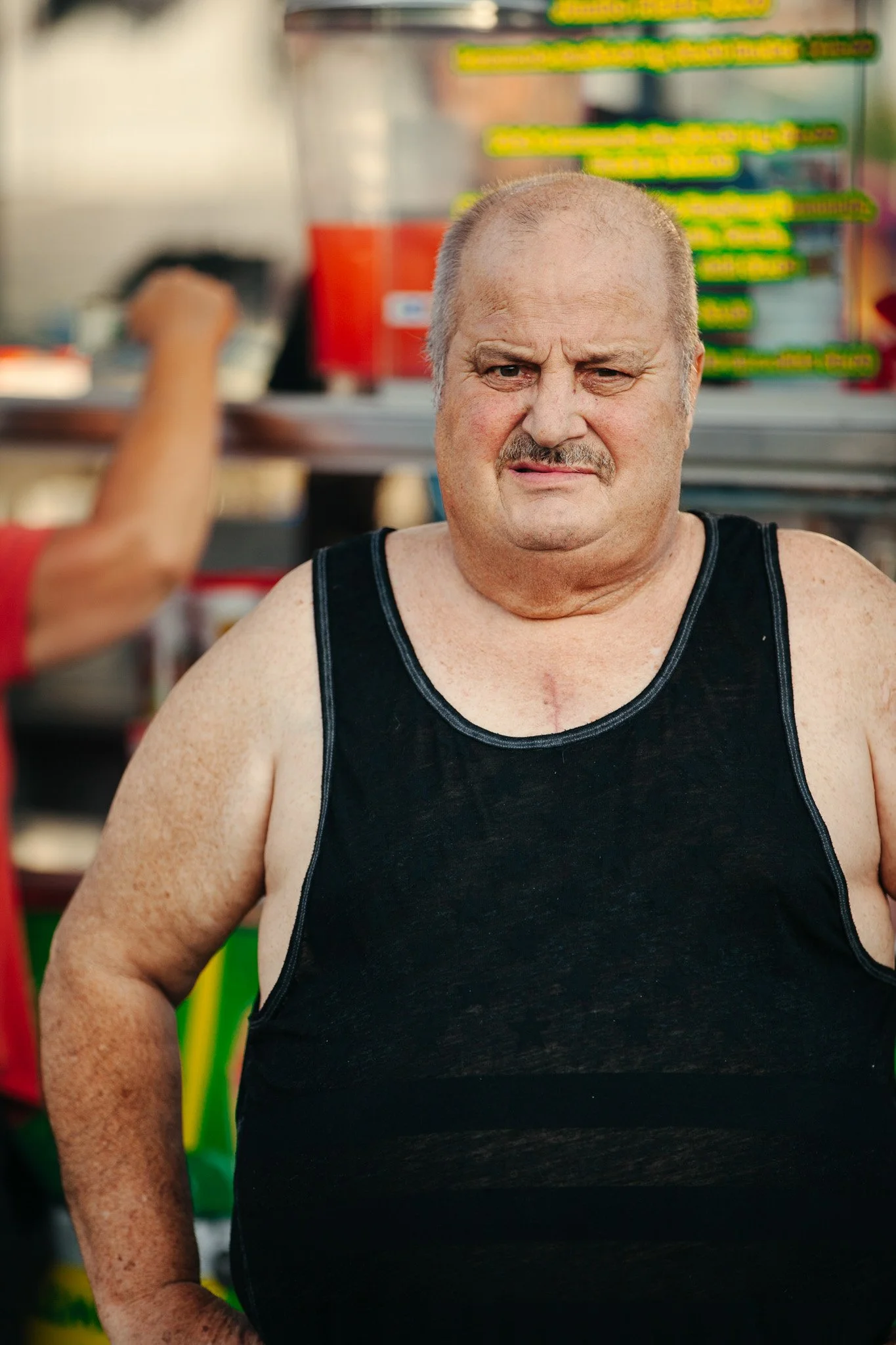 A middle-aged man with a serious expression, wearing a black sleeveless shirt, in a store with colorful shelves and blurred products in the background.