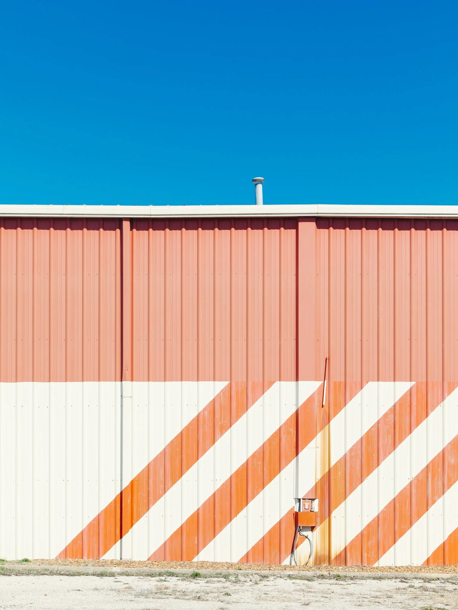 A building with metal siding painted in pink, white, and orange colors, with diagonal orange and white stripes near the bottom. The top of the building has a white trim, and a chimney protrudes above the roofline. There is a concrete sidewalk and dir