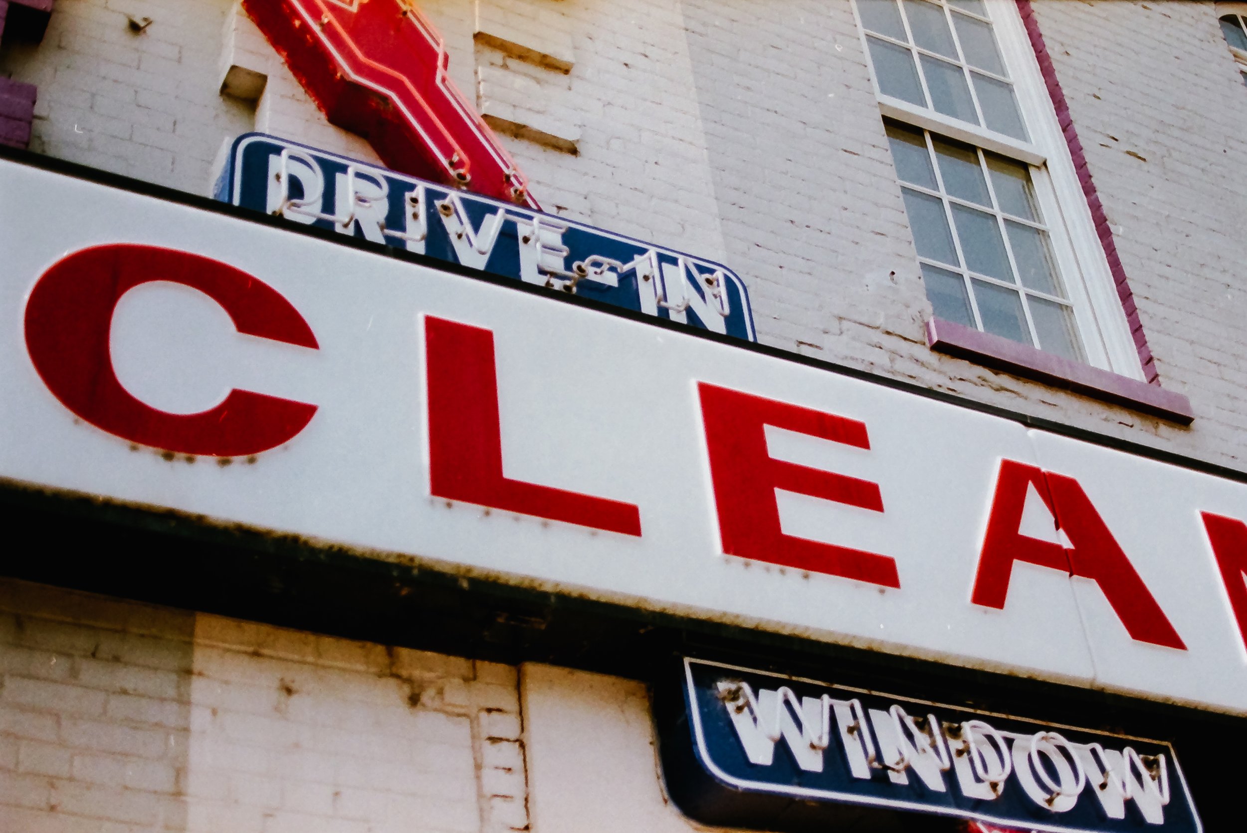 Close-up of a vintage neon sign reading 'Drive-In' mounted on a brick building with windows and a partial view of a yellow sign below.