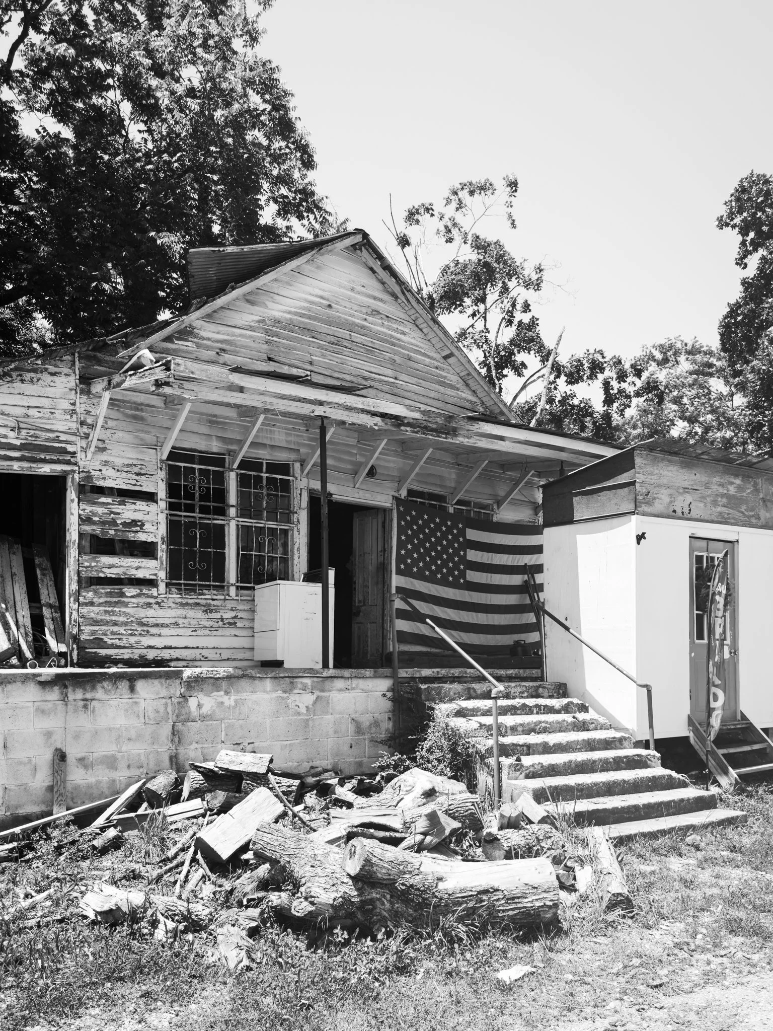 Old, weathered house with a flag hanging on the porch, surrounded by trees and a pile of chopped firewood in front.