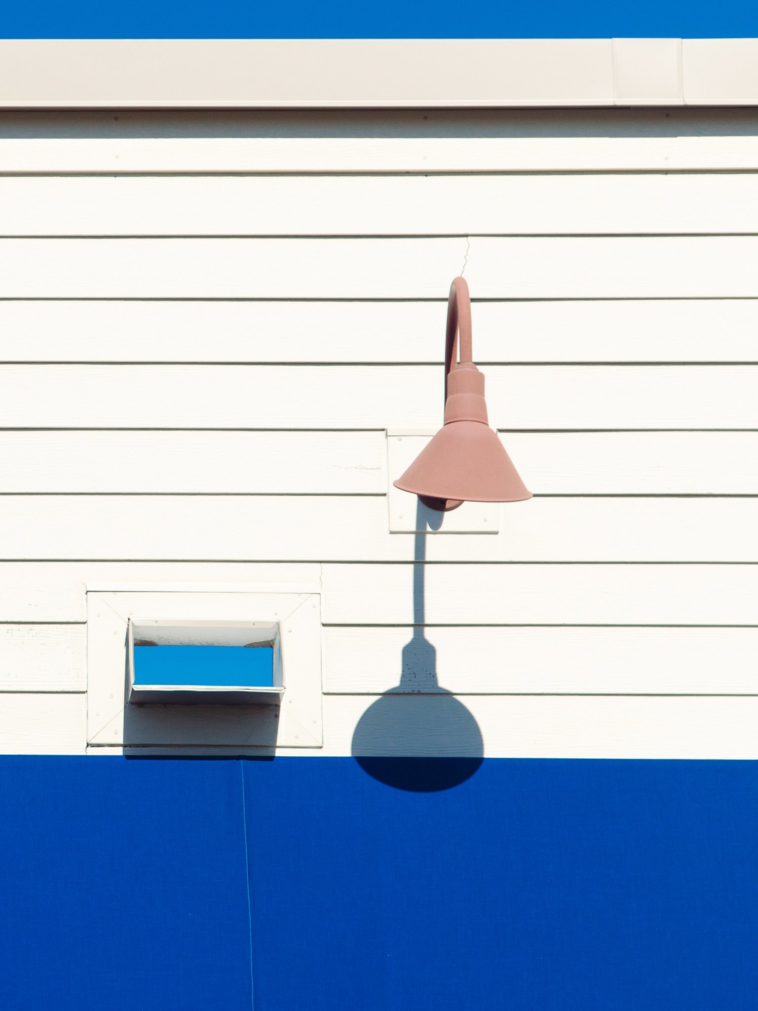 Exterior white house wall with a small window and a pink outdoor cone-shaped light fixture casting a shadow on the wall.