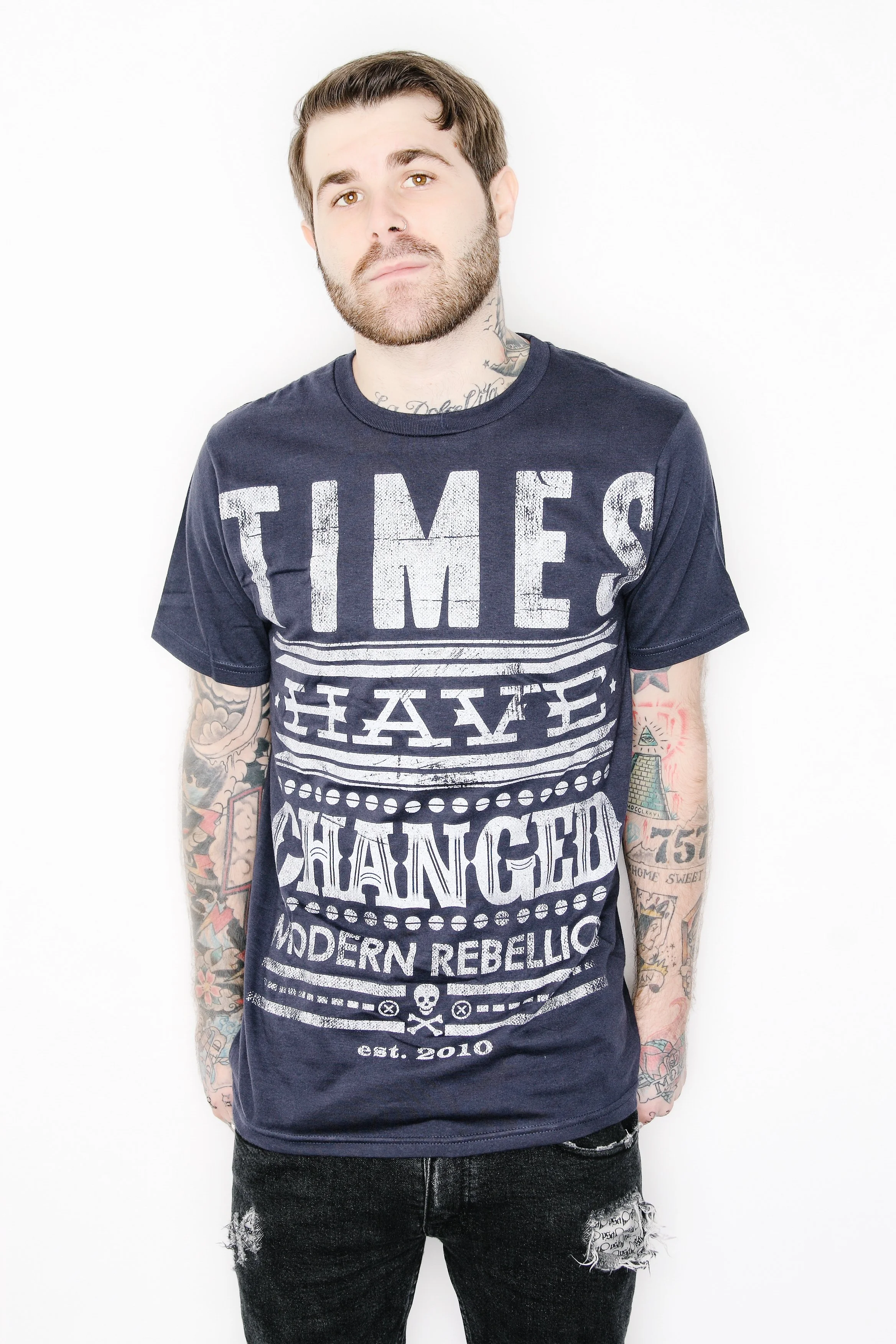 A young man with brown hair, a beard, and tattoos on his arms and neck, wearing a dark blue graphic T-shirt and distressed black jeans, standing against a white background.