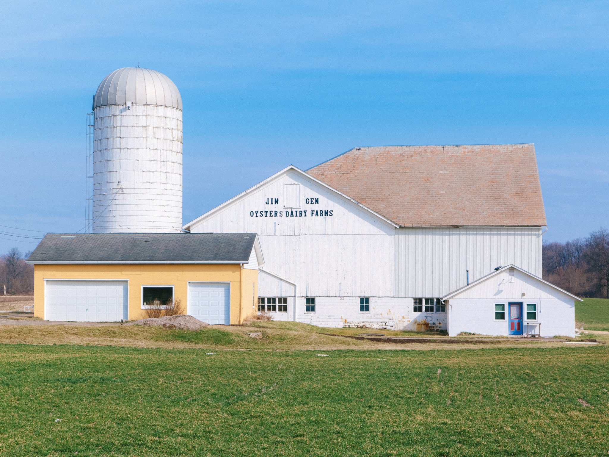 A farm building with a white barn and a yellow garage, with a large white silo to the left. The barn has the words "Jim Genn Oysters Dairy Farms" written on it. The sky is clear and blue, and there is green grass in the foreground.