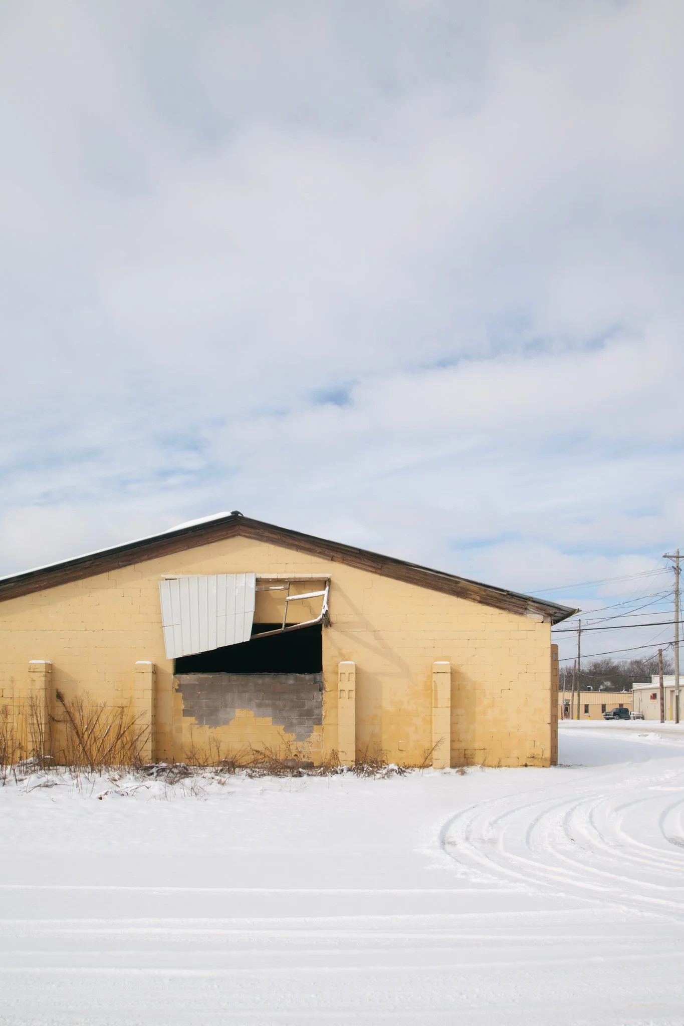 A yellow house with damage on the front, missing a window, and rotting snow in the foreground. The sky is cloudy.