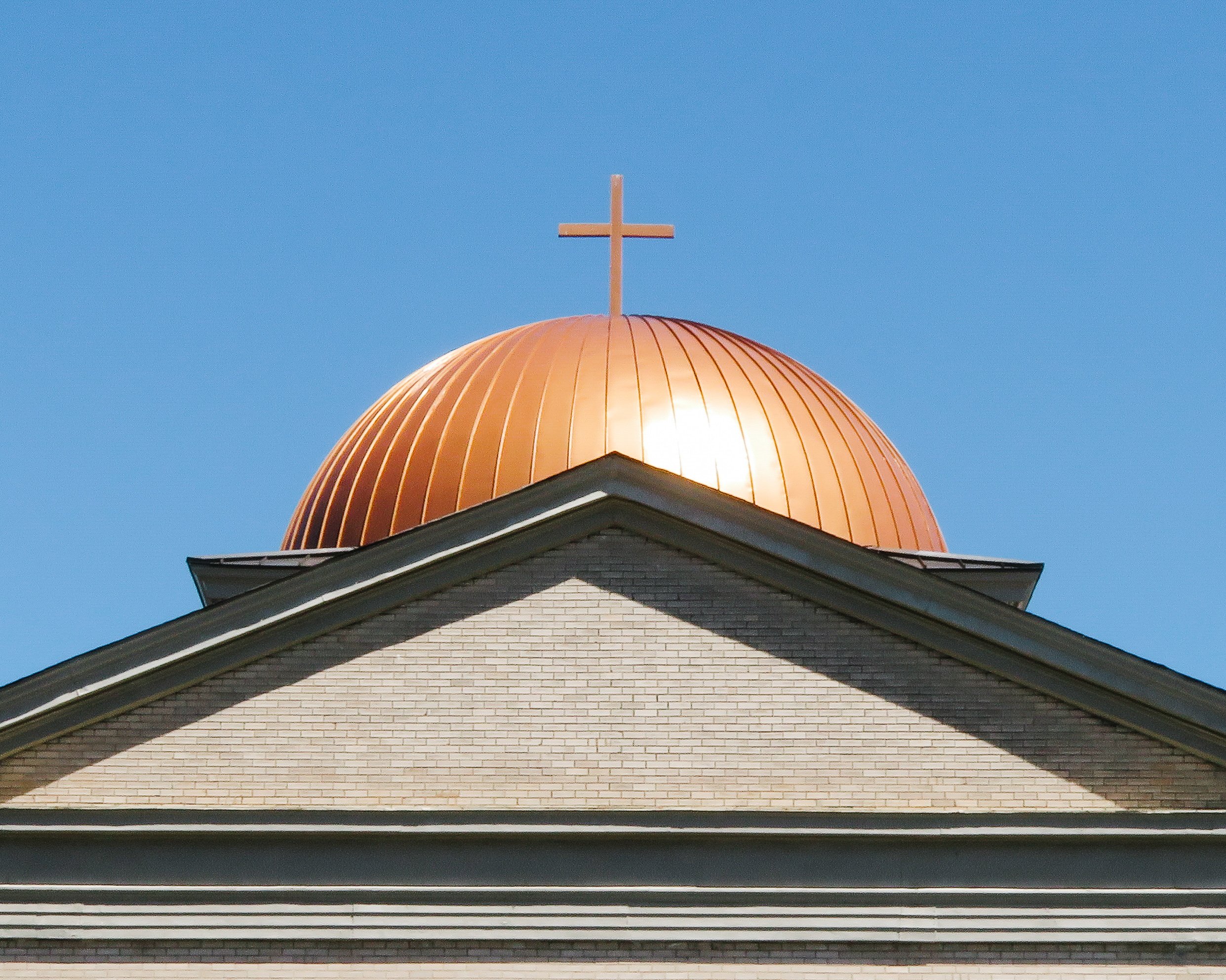 The image shows the top portion of a church with a white brick facade, a rounded copper-colored dome, and a cross on top of the dome, set against a clear blue sky.