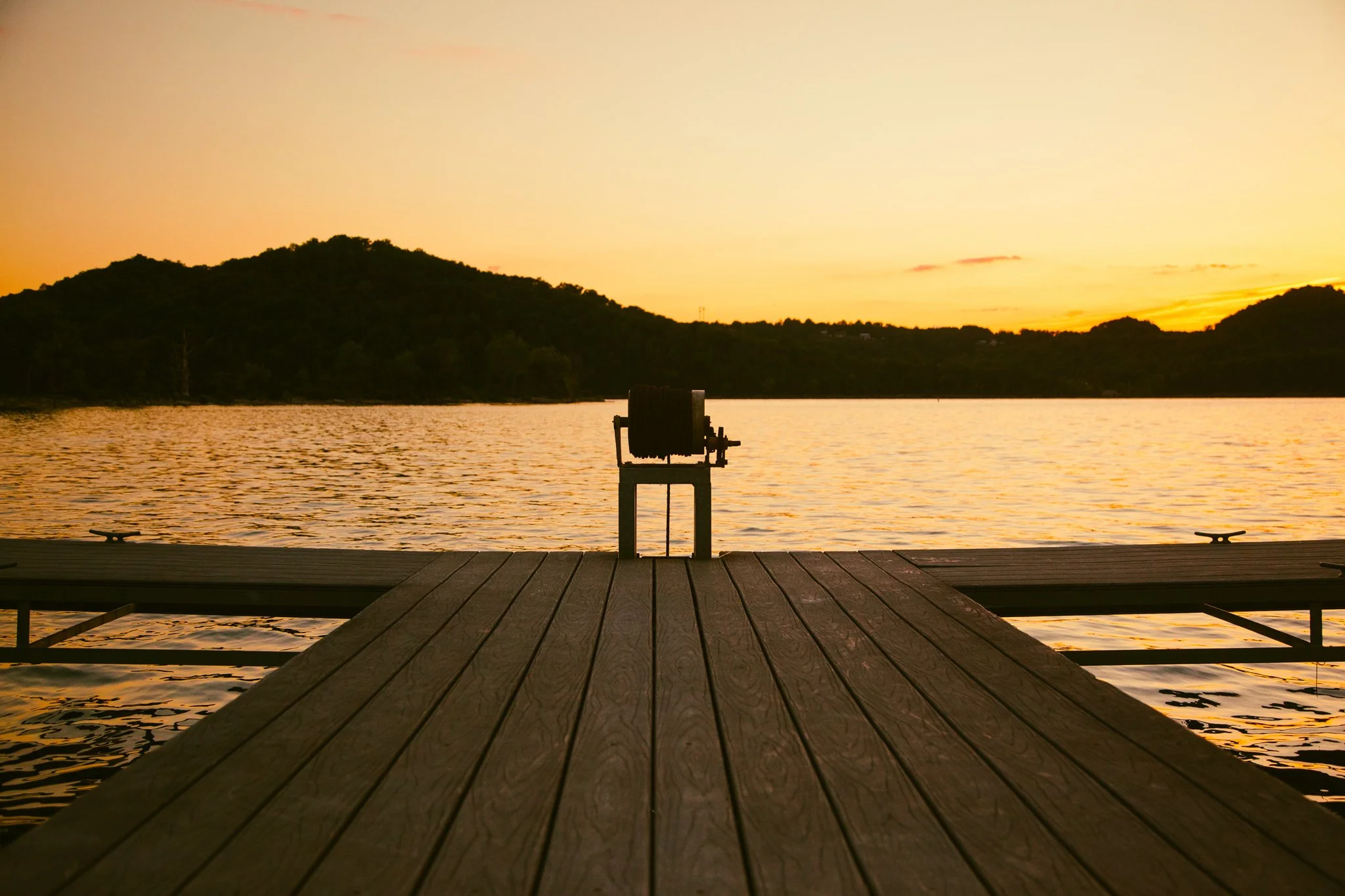 A wooden dock extending over a body of water with hills in the background during sunset.
