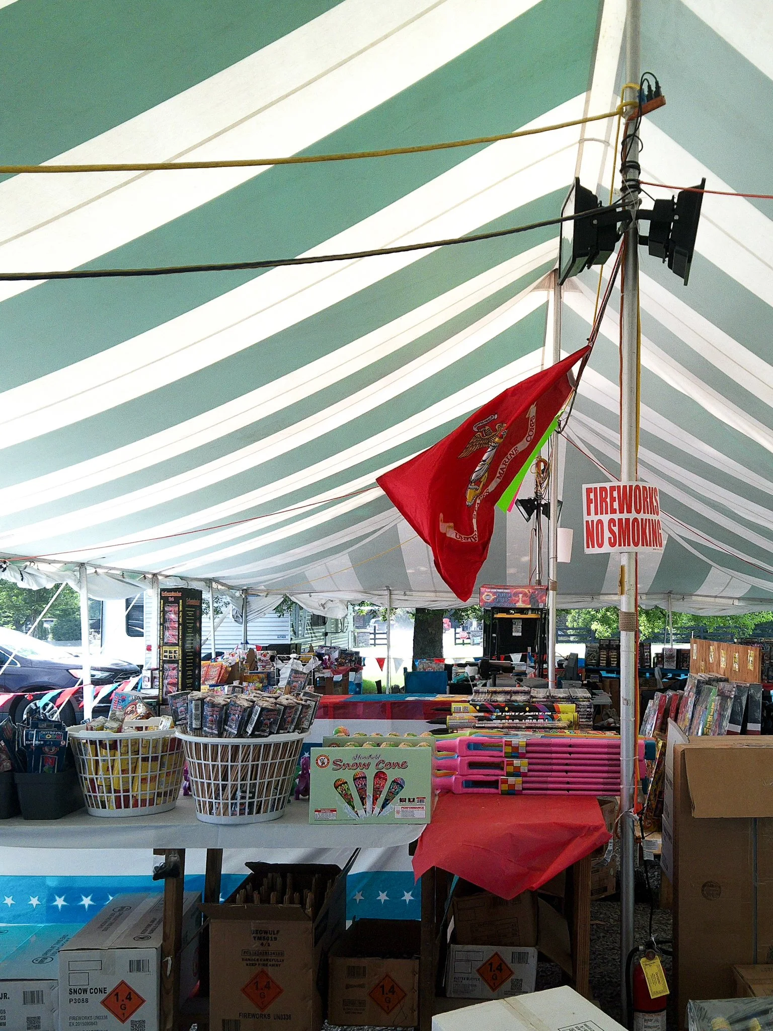 A marketplace booth under a green and white striped canopy, with various items like books and toys on display, and boxes labeled 'fireworks' with hazard symbols.