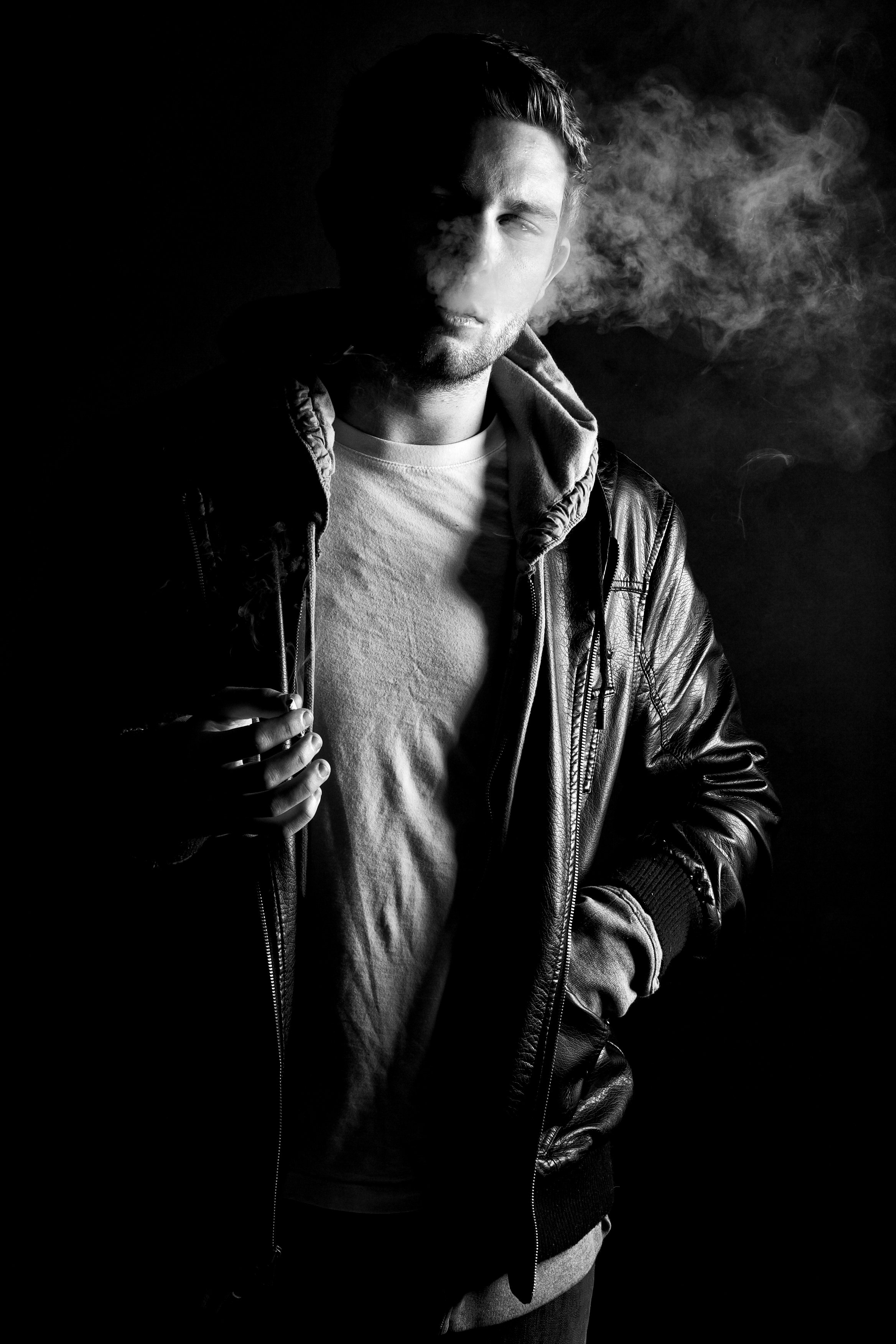 Black and white portrait of a young man with short hair, wearing a leather jacket over a hoodie and T-shirt, holding a cigarette in his right hand.
