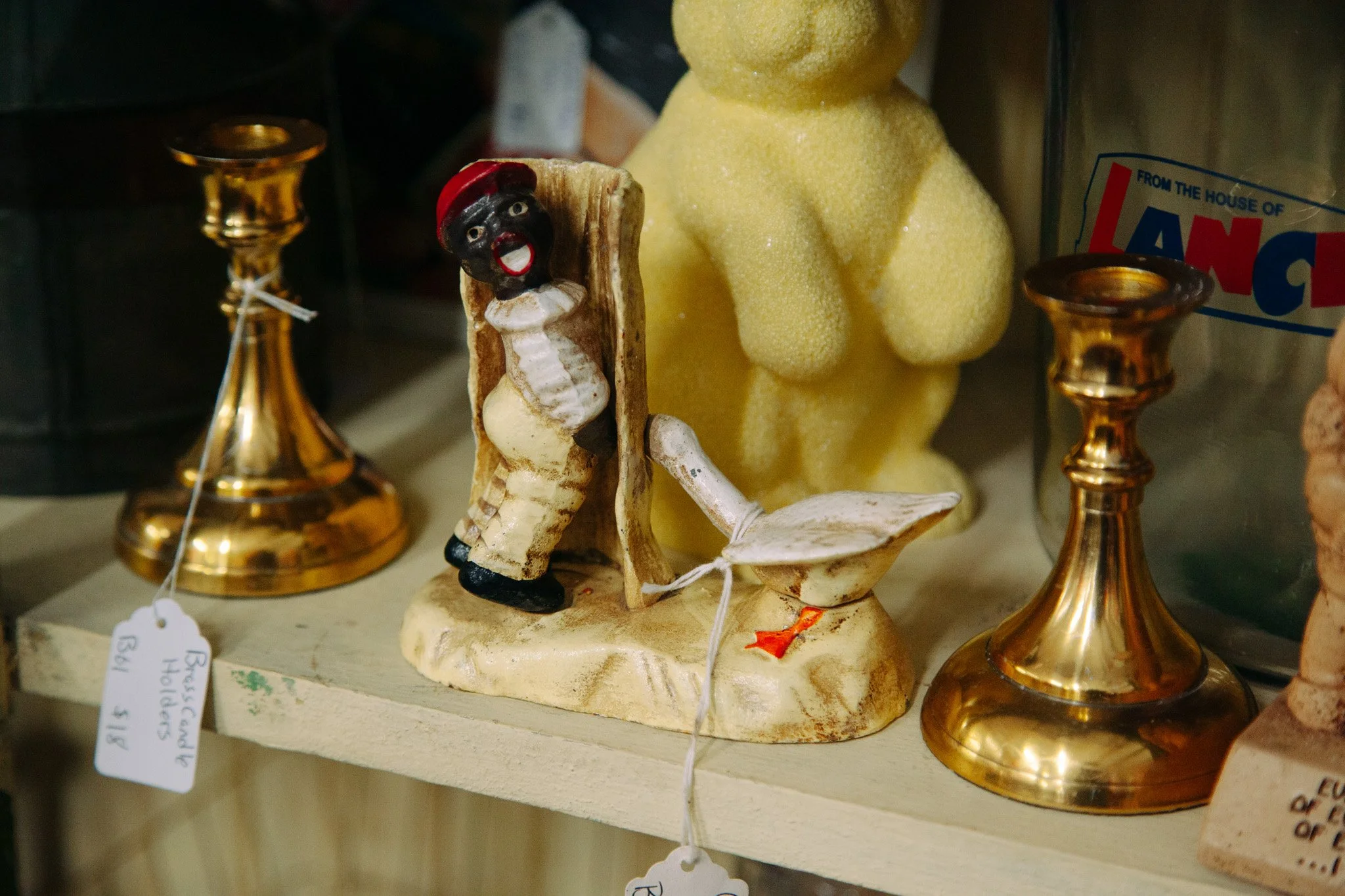 A small figurine of a person with dark skin, white clothing, and a red hat, standing next to two gold-colored candle holders. Behind the figurine is a yellow textured Christmas tree ornament and a sign partially visible on the right side.