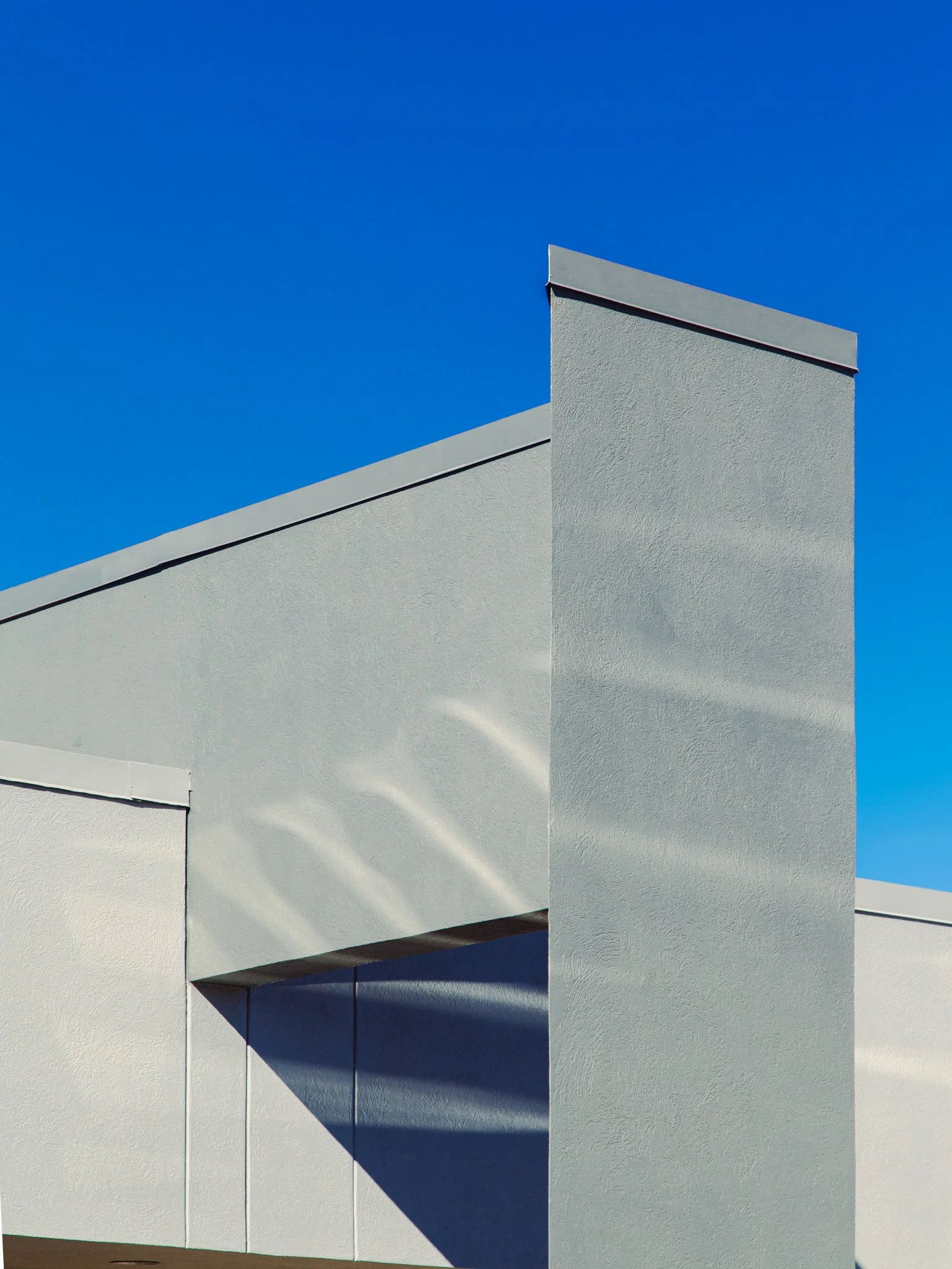 Close-up of a modern white building with textured exterior walls and sharp angles, set against a clear blue sky, with shadows cast on the walls.