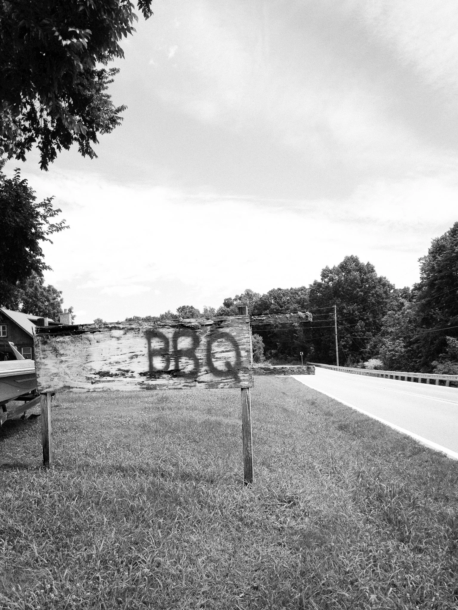 A black-and-white photo of a rural scene with a grassy area in the foreground, a weathered wooden sign with graffiti, trees on both sides, and a curving road in the distance under a cloudy sky.