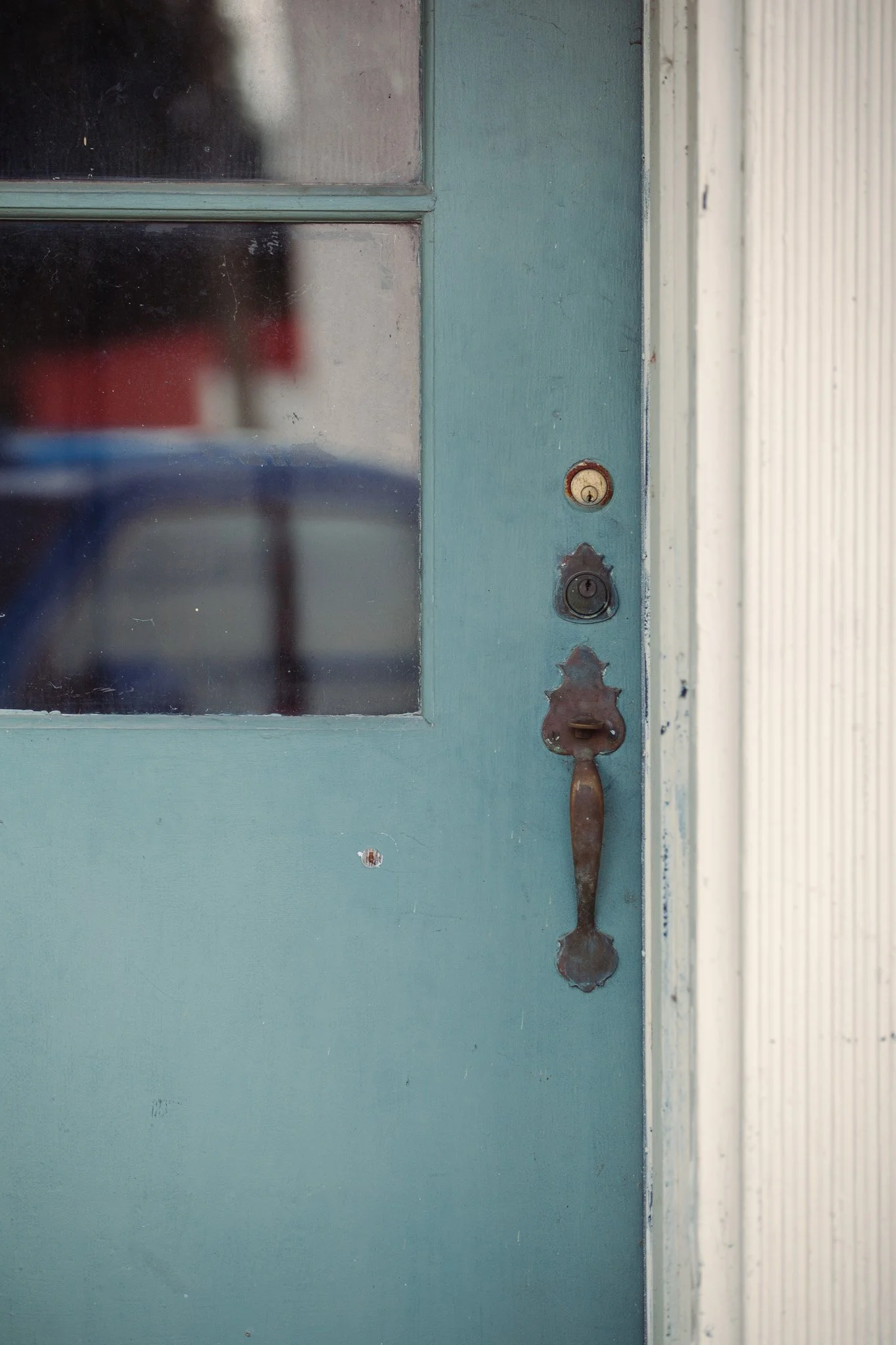 Close-up of an old, weathered blue door with a glass window. The door has a vintage, rusty door handle and a keyhole with an aged escutcheon plate. The window is dirty and shows a blurred reflection of an outdoor scene.
