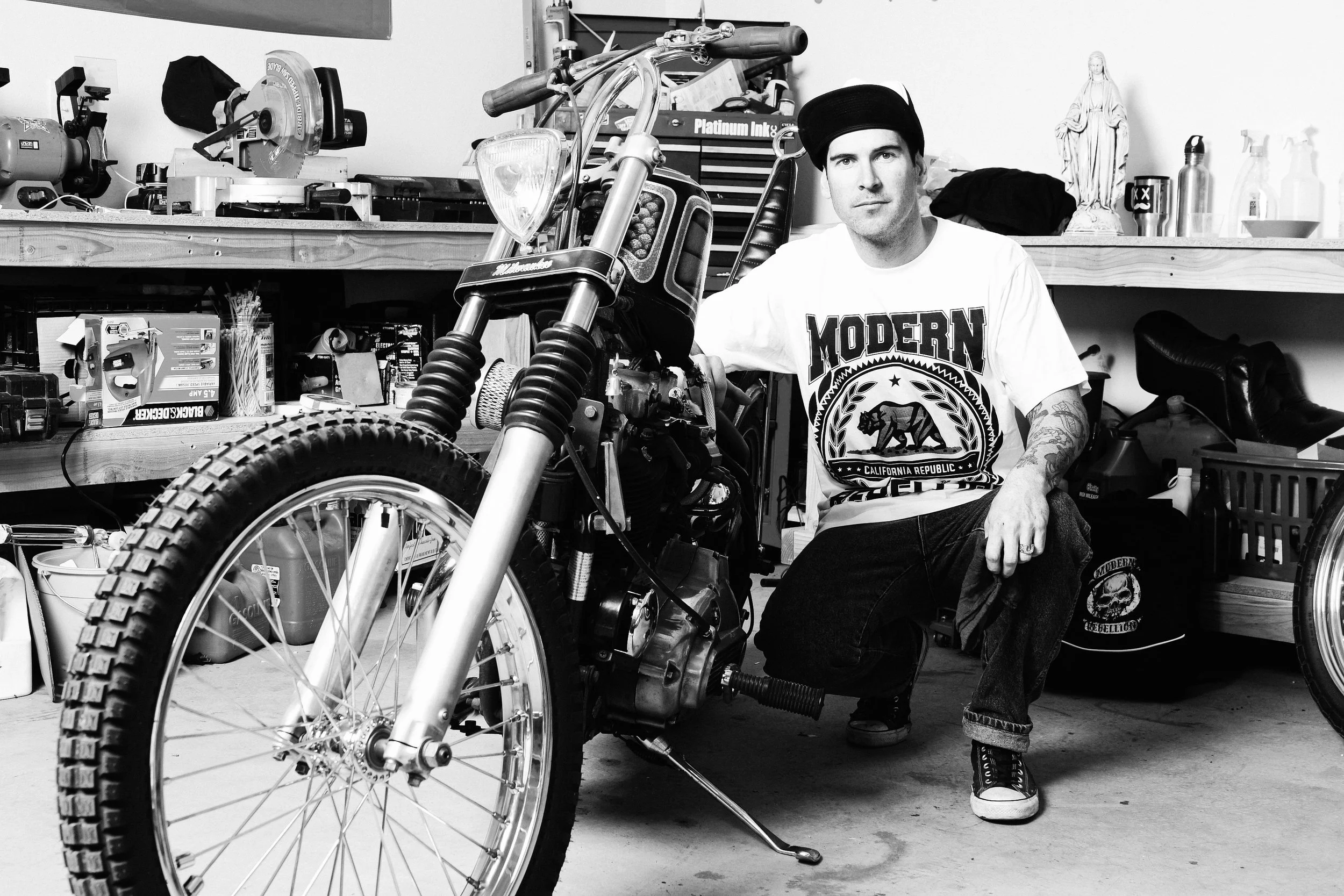 A man wearing a black cap, white T-shirt with 'Modern' logo, and jeans, sitting in a garage or workshop beside a disassembled motorcycle. The background includes tools, a statue, and various bottles and boxes on shelves.