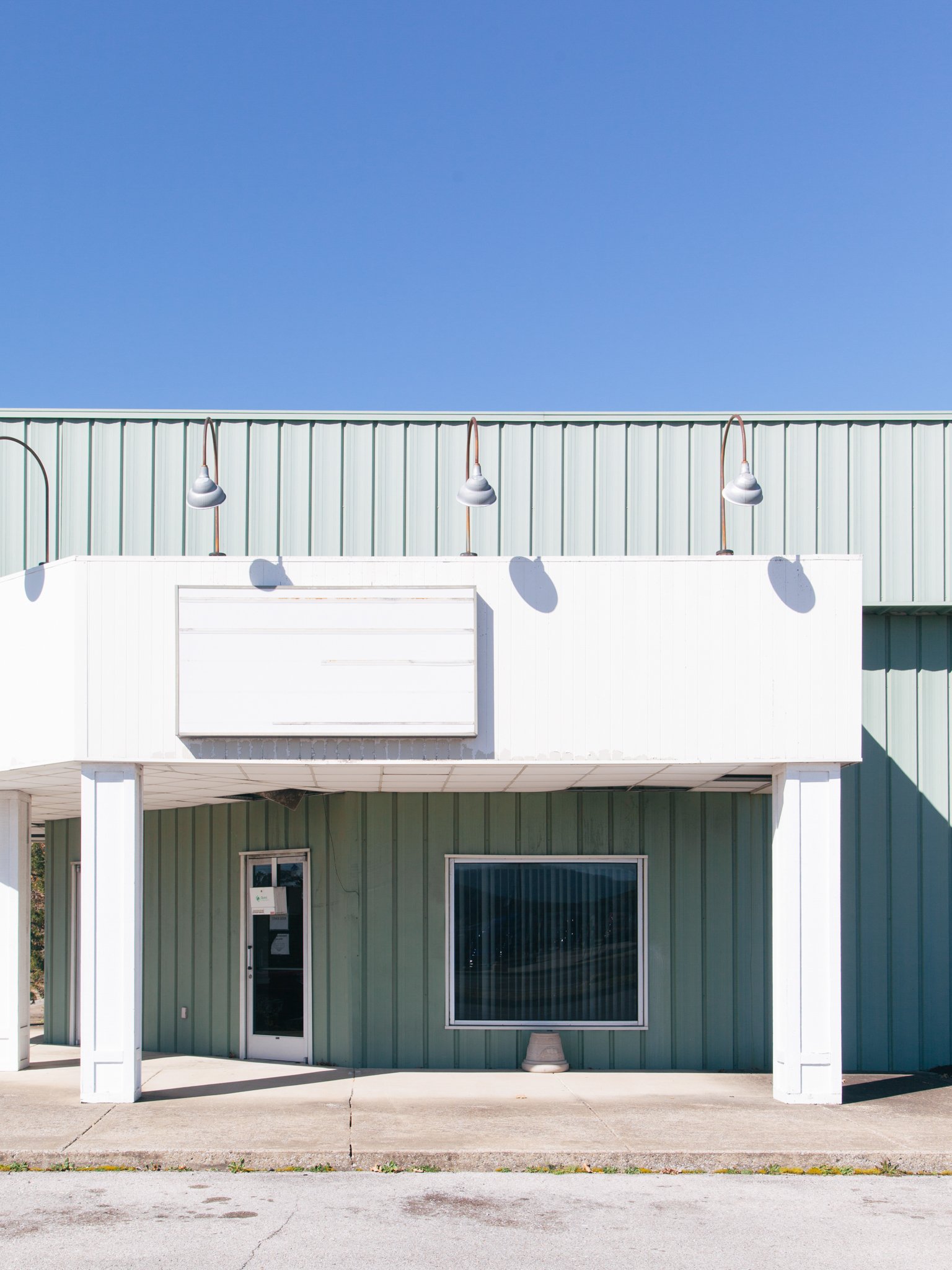Front view of a commercial building with a green exterior wall, white trim, large window, and a door, under a clear blue sky. There are four outdoor lights mounted on the wall above the entrance.