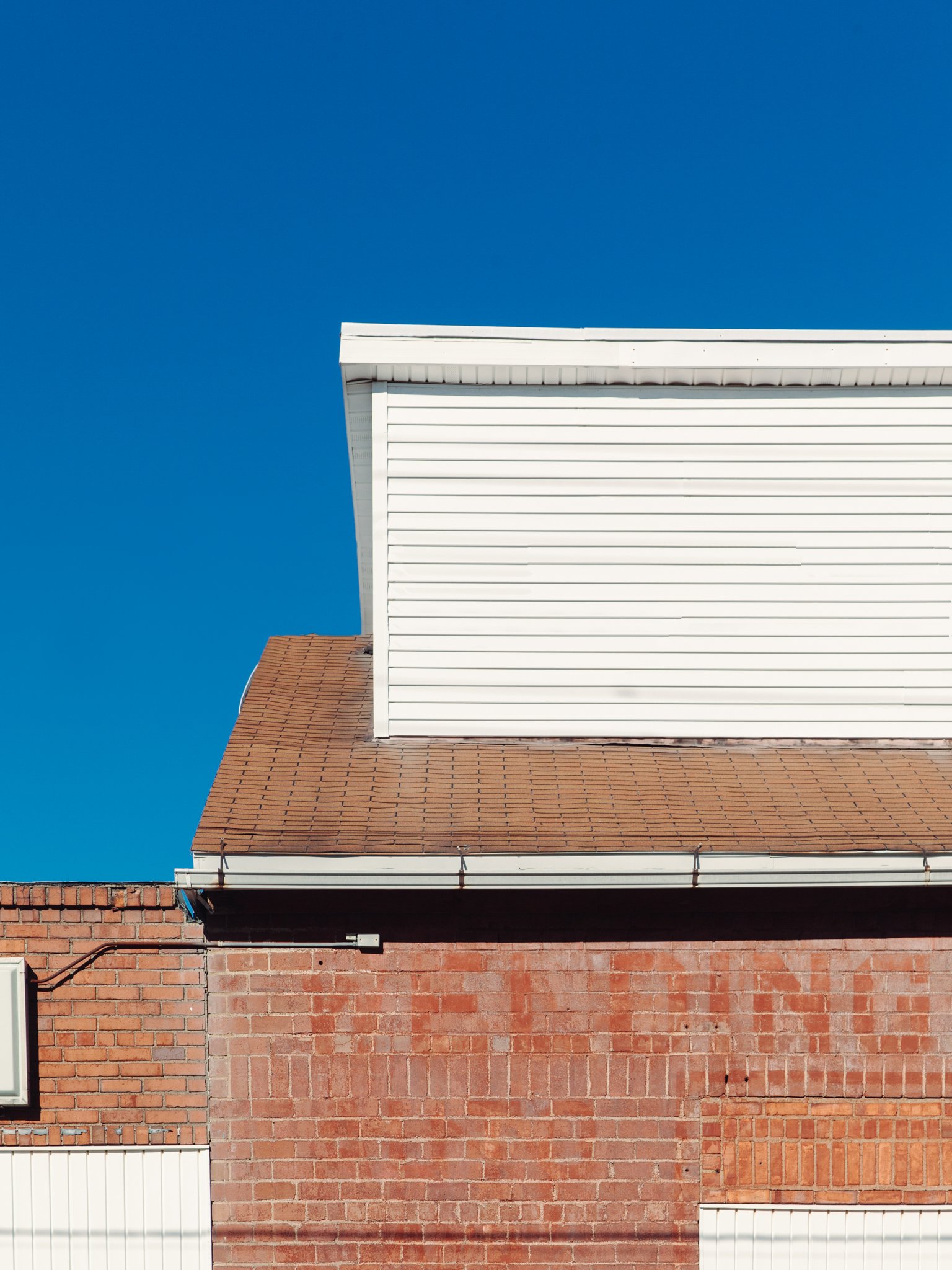The image shows the corner of a building with a brick wall at the bottom and a section with white siding on the upper part. The roof has brown shingles, and the sky is clear and blue.