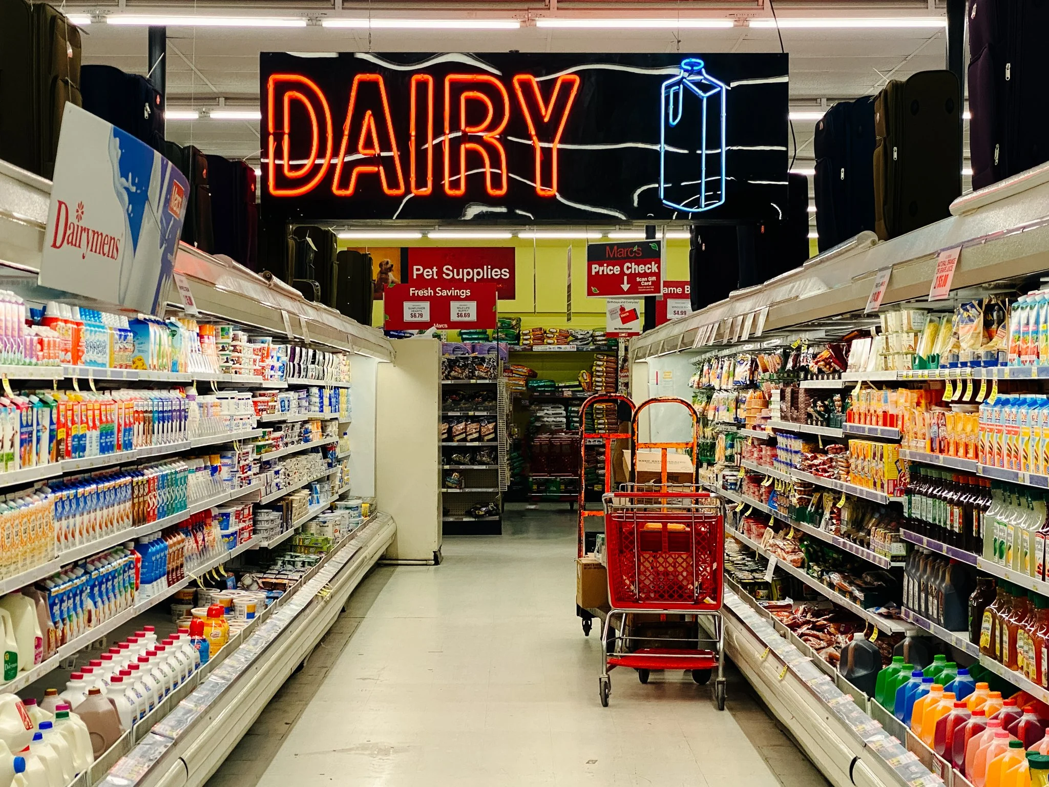 Store aisle with milk and juice bottles on the left, cleaning products on the right, and a red shopping cart in the center. Neon sign reads 'DAIRY' with a milk carton icon above.