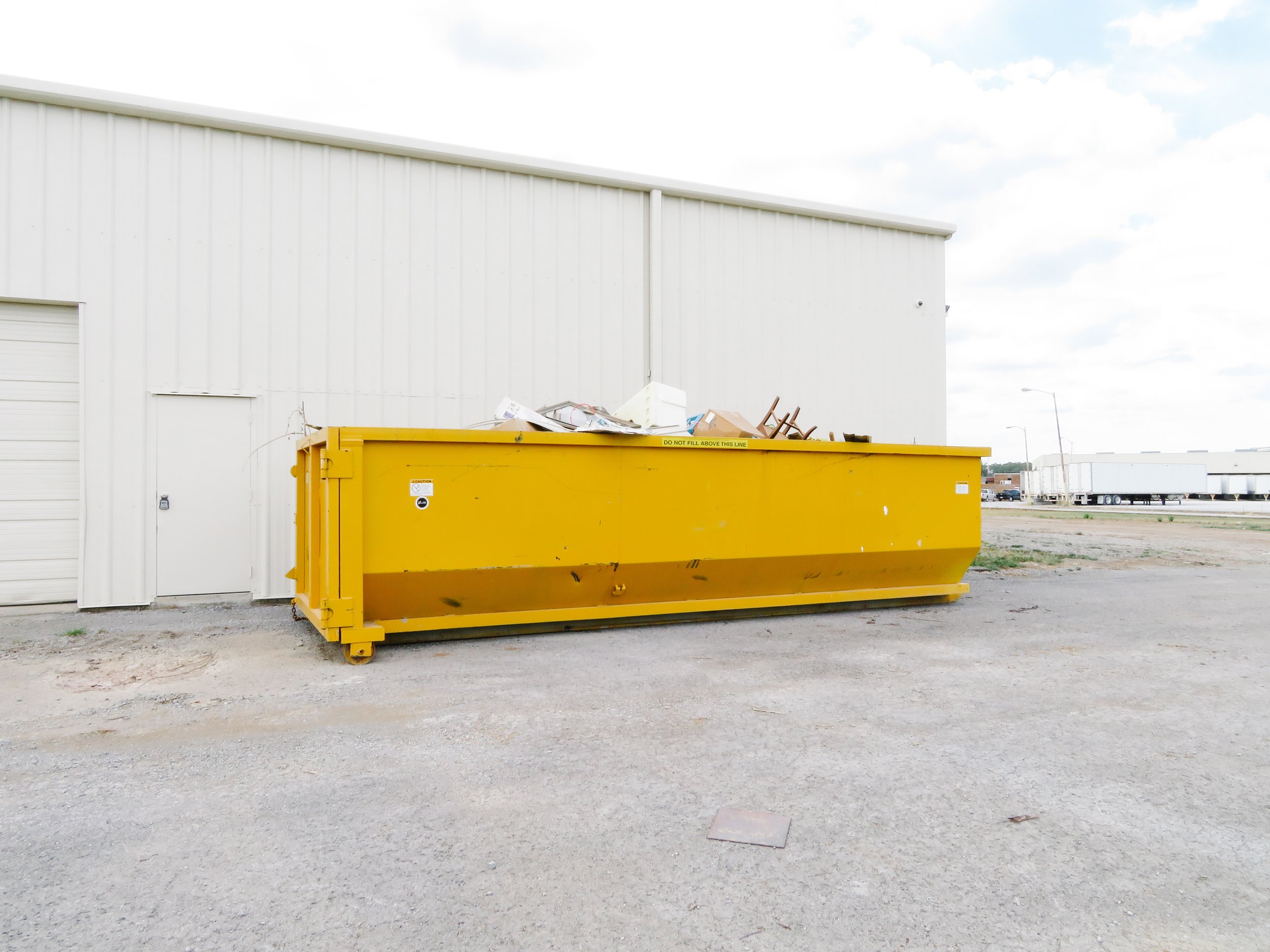 A large yellow dumpster filled with trash such as cardboard, plastic, and paper, positioned outside a white industrial building with a rolled garage door and a sky with scattered clouds in the background.