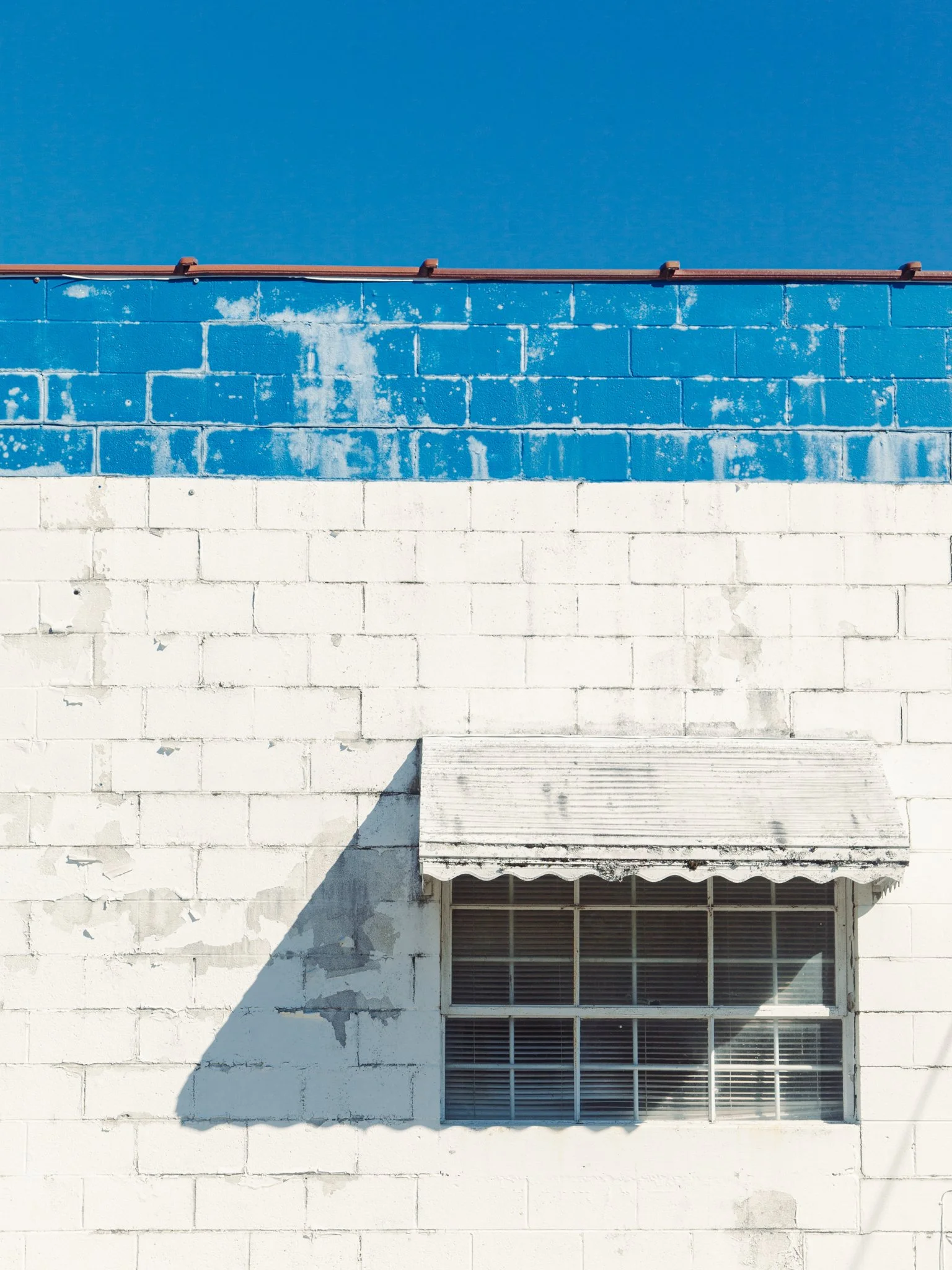 The image shows a section of a weathered white brick building with a large window and a closed metal shutter awning. The brick wall has peeling paint and signs of wear, with a shadow cast across part of the wall.