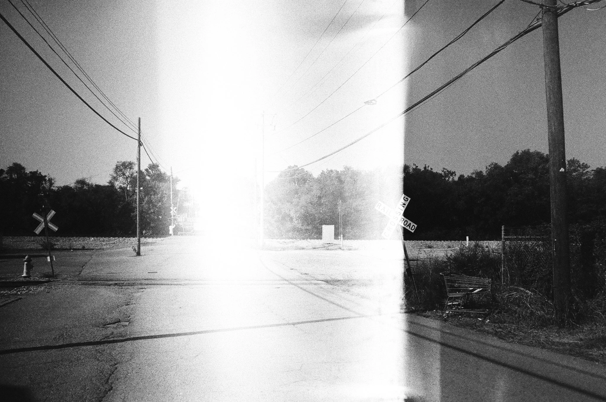 Black and white photo of an empty intersection with railroad crossing signs, utility poles, and power lines, some trees in the background, and sunlight creating glare in the center.
