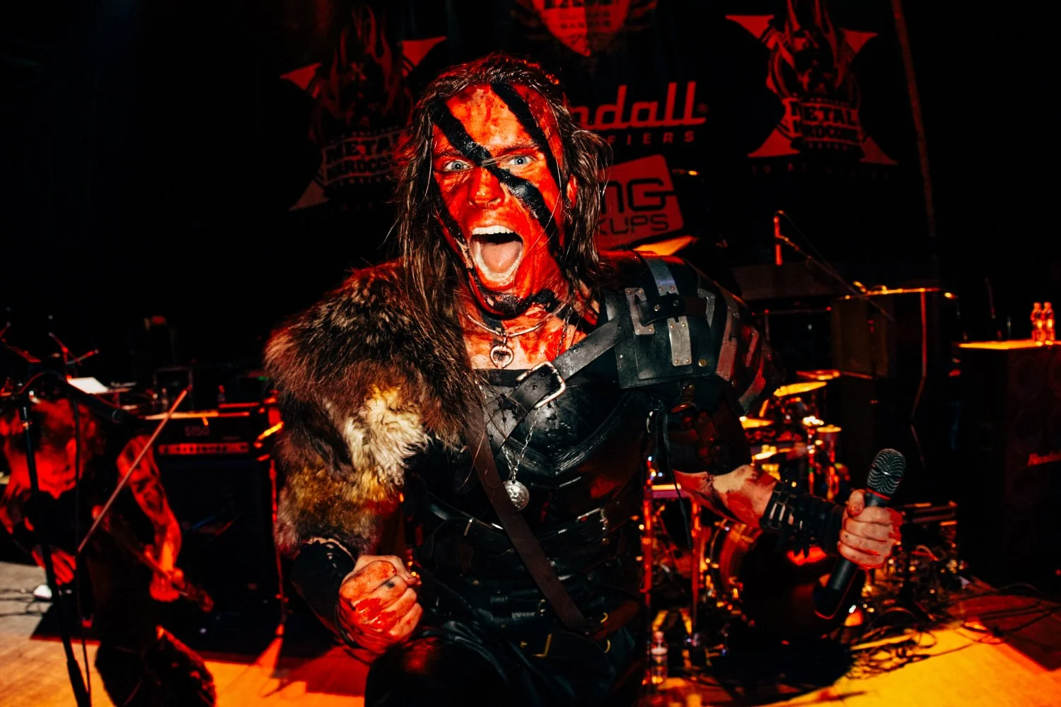 A musician in black leather and fur clothing, with face painted red and black, screaming passionately on stage holding a microphone, with intense stage lighting and a concert background.