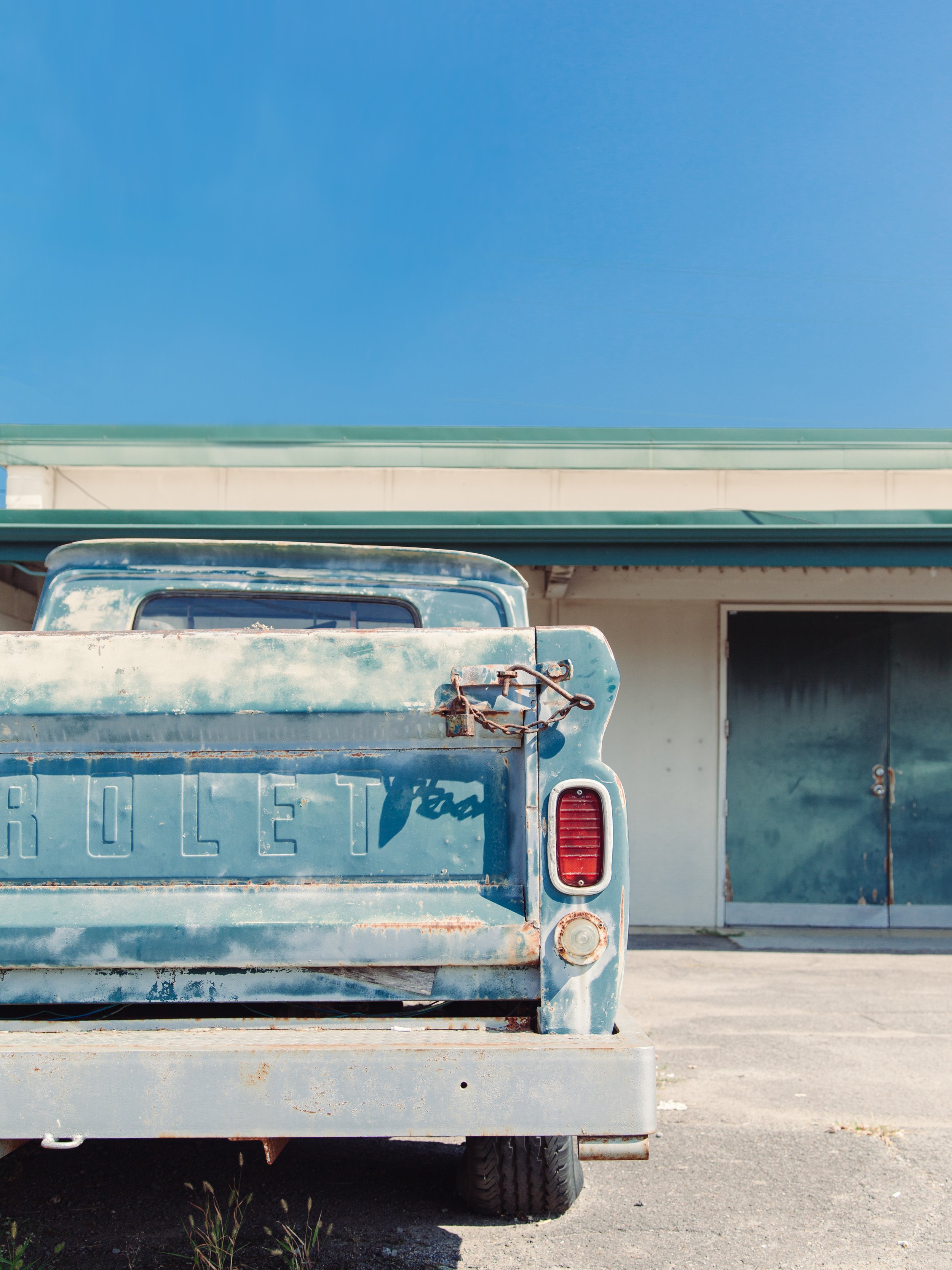 Close-up of the rear of a vintage blue pickup truck with rust and weathering, parked on a concrete surface in front of a building with a green roof and a blue sky background.