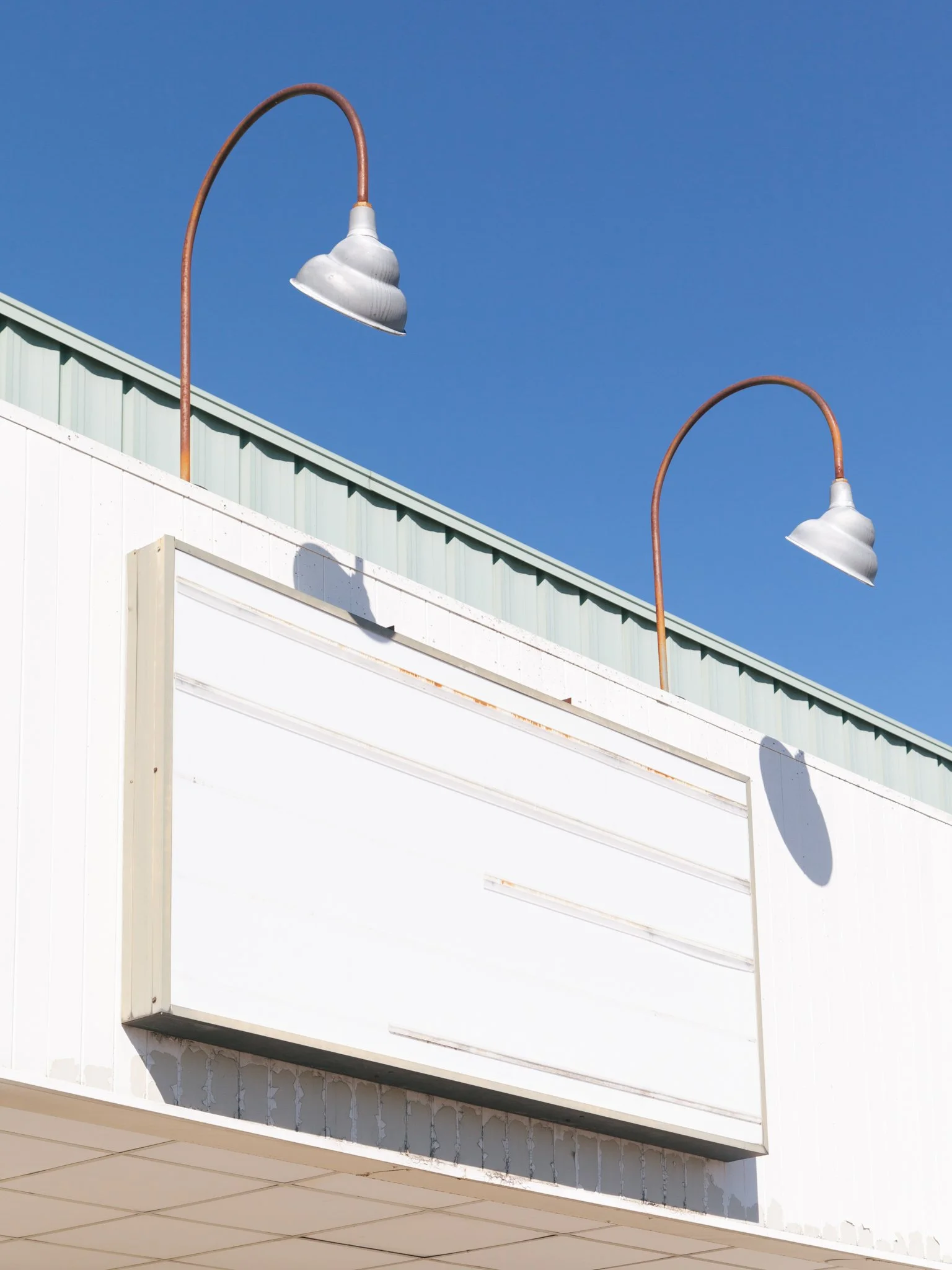 Two modern outdoor lamps with curved, rusty metal poles mounted on a building facade, set against a clear blue sky.