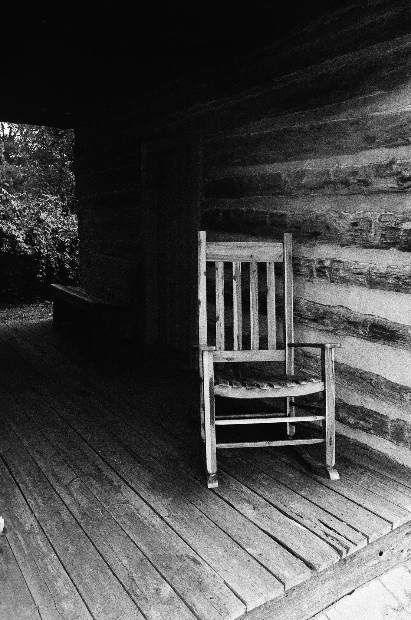 A wooden porch with curved flooring, a rustic log wall, a wooden chair, and a corner of a table, with bushes and trees outside the porch.