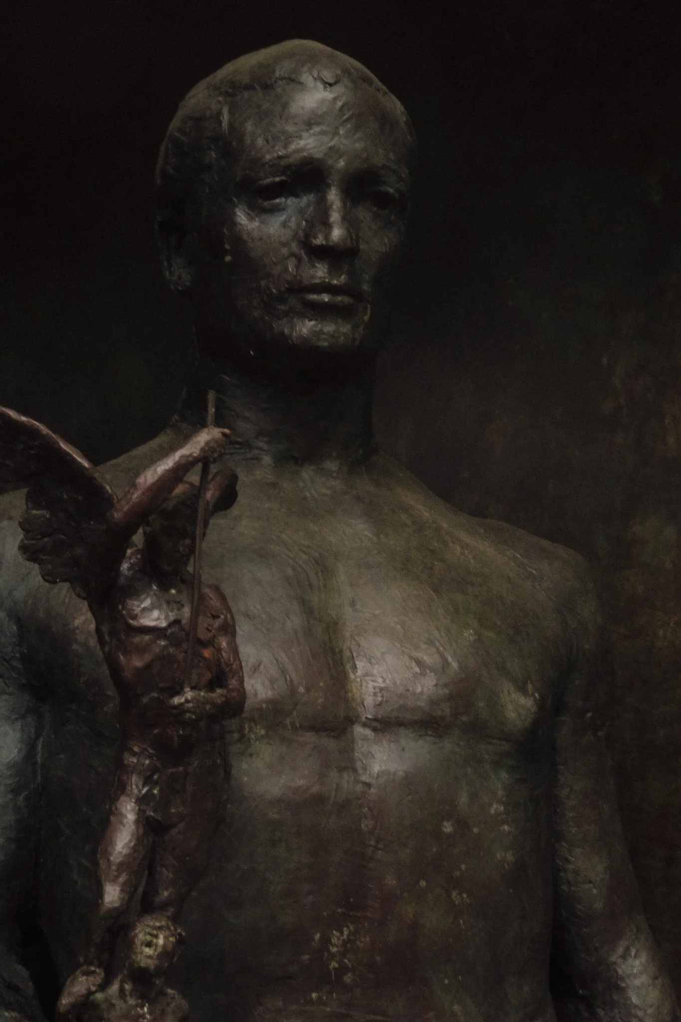 A bronze sculpture of a woman with a solemn expression, holding a small object close to her face, set against a dark background.