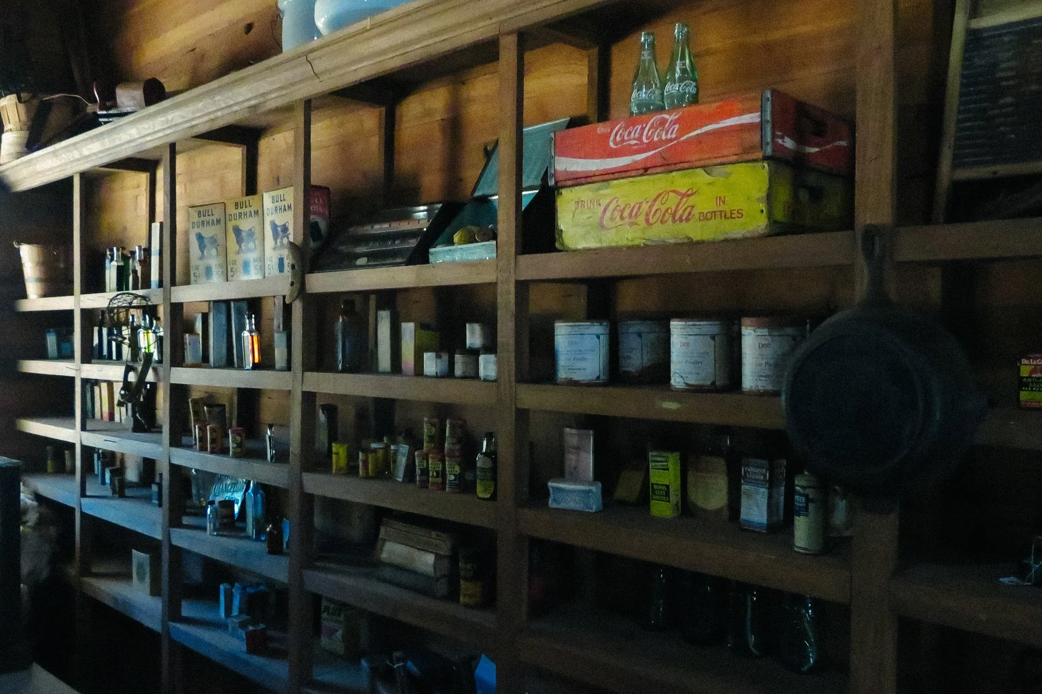 Wooden shelves stocked with vintage Coca-Cola boxes, glass bottles, and various canned and bottled beverages in a dimly lit room.