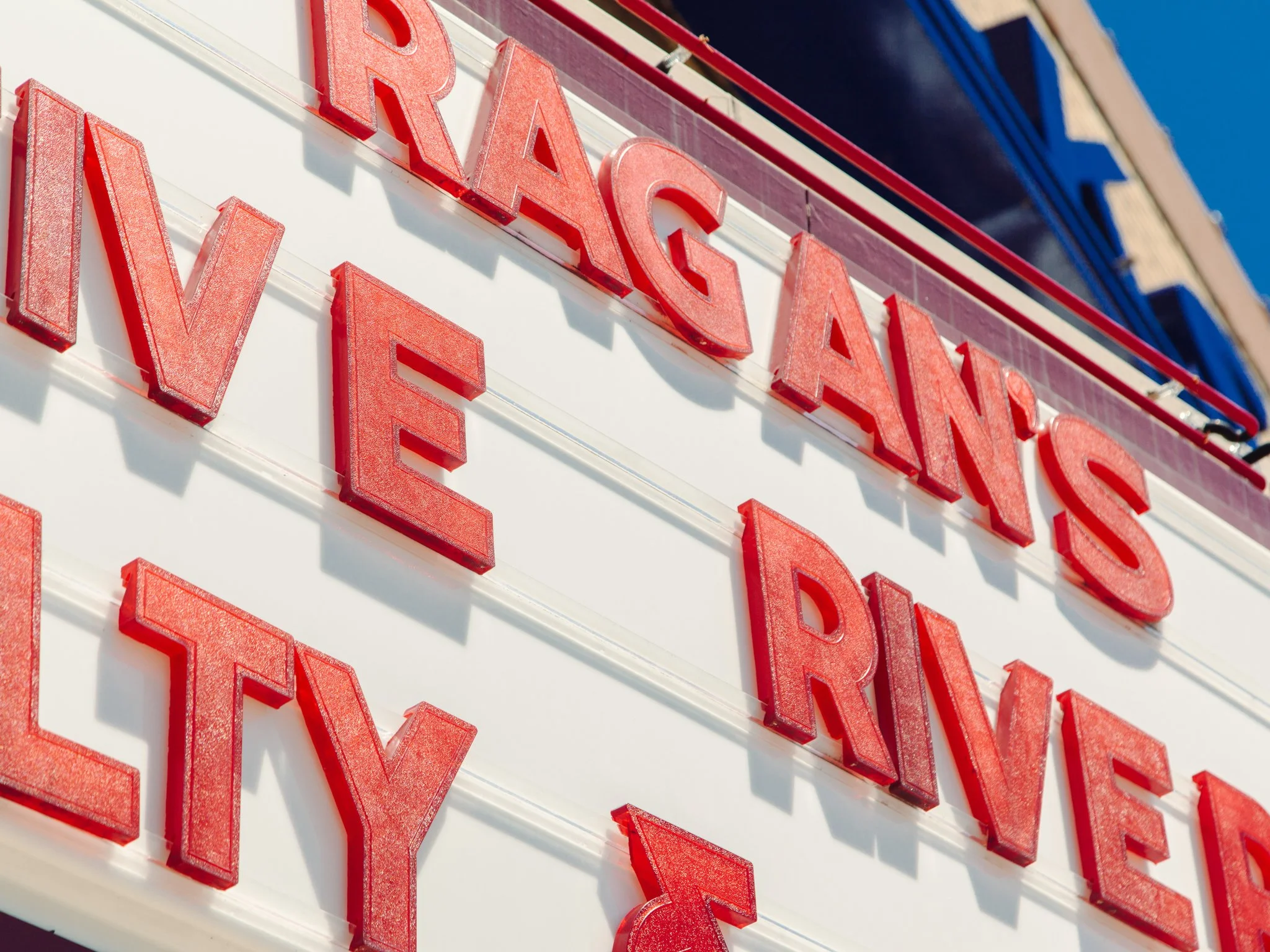 Close-up of a red neon sign on a white building with the word 'PRACT' visible, with a blue sky in the background.