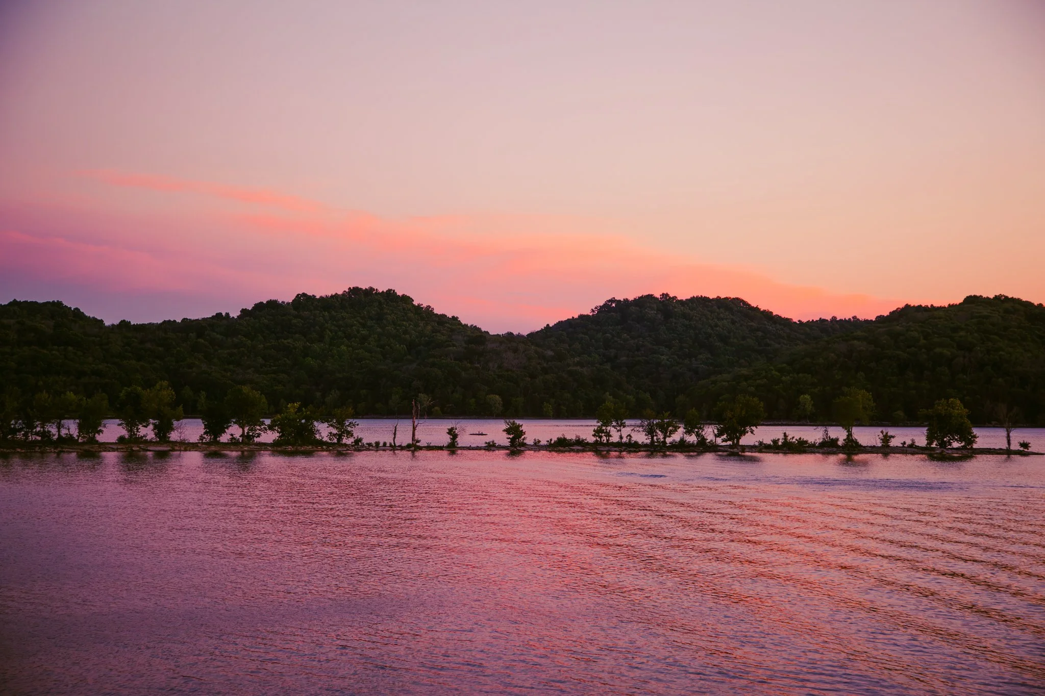Sunset over a river with a line of trees and hills in the background, displaying pink and purple colors in the sky.