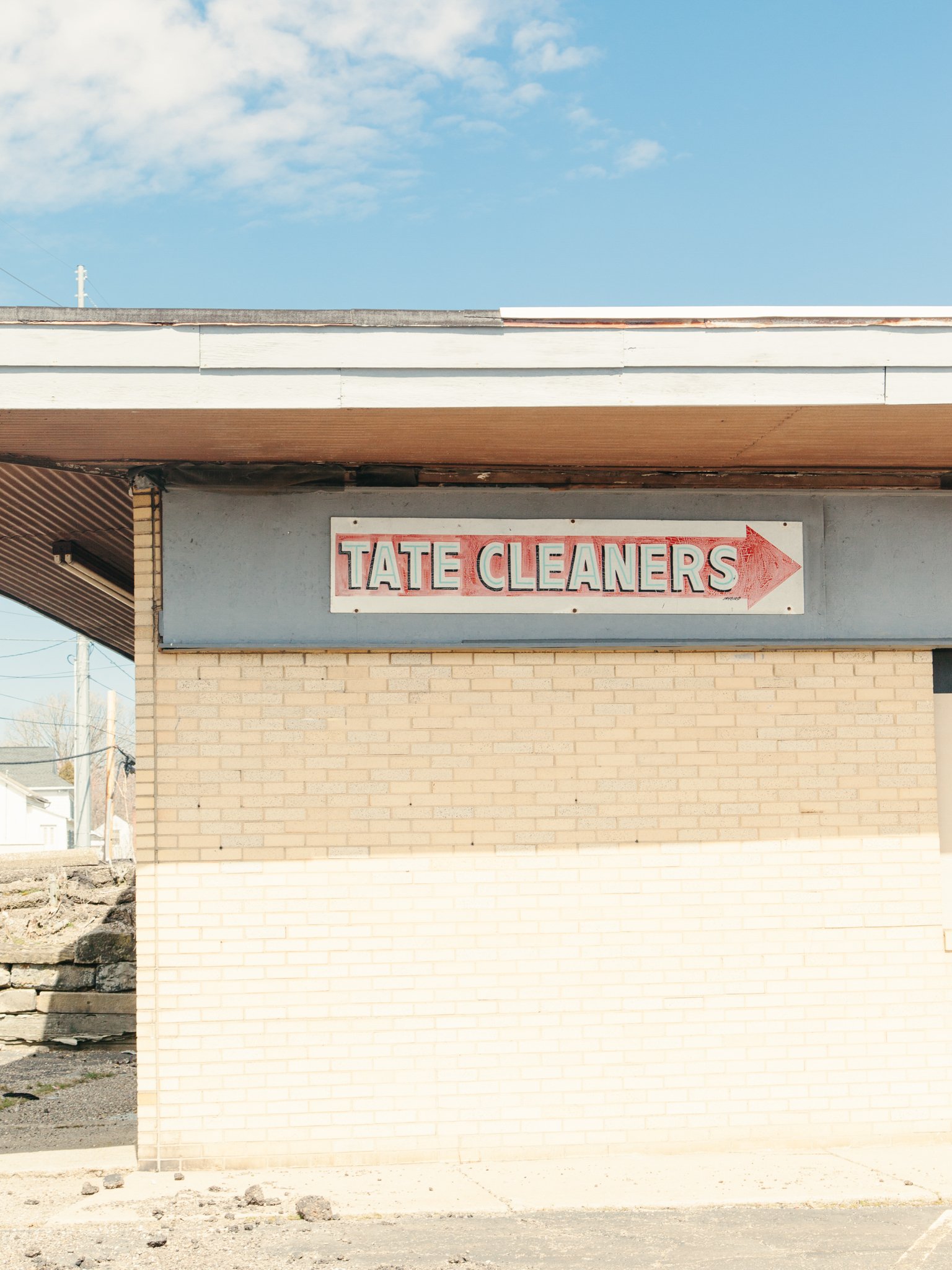 A beige brick building with a sign that reads 'TATE CLEANERS' with an arrow pointing to the right, set against a blue sky with some clouds.