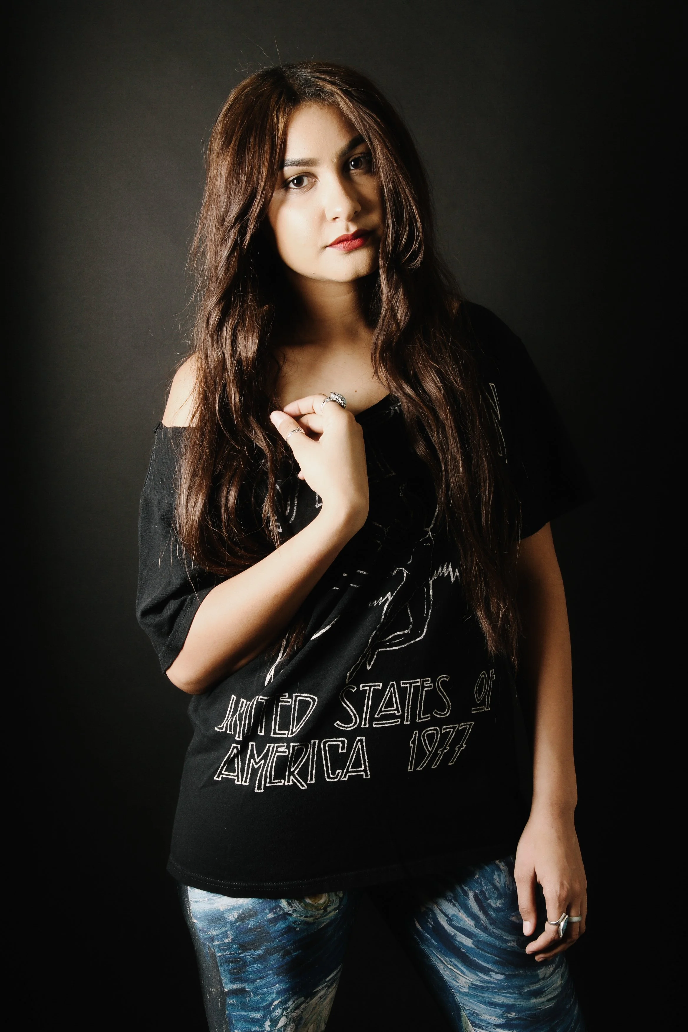 Young woman with long wavy brown hair wearing a black graphic t-shirt and vibrant blue and white patterned leggings, standing against a dark background.