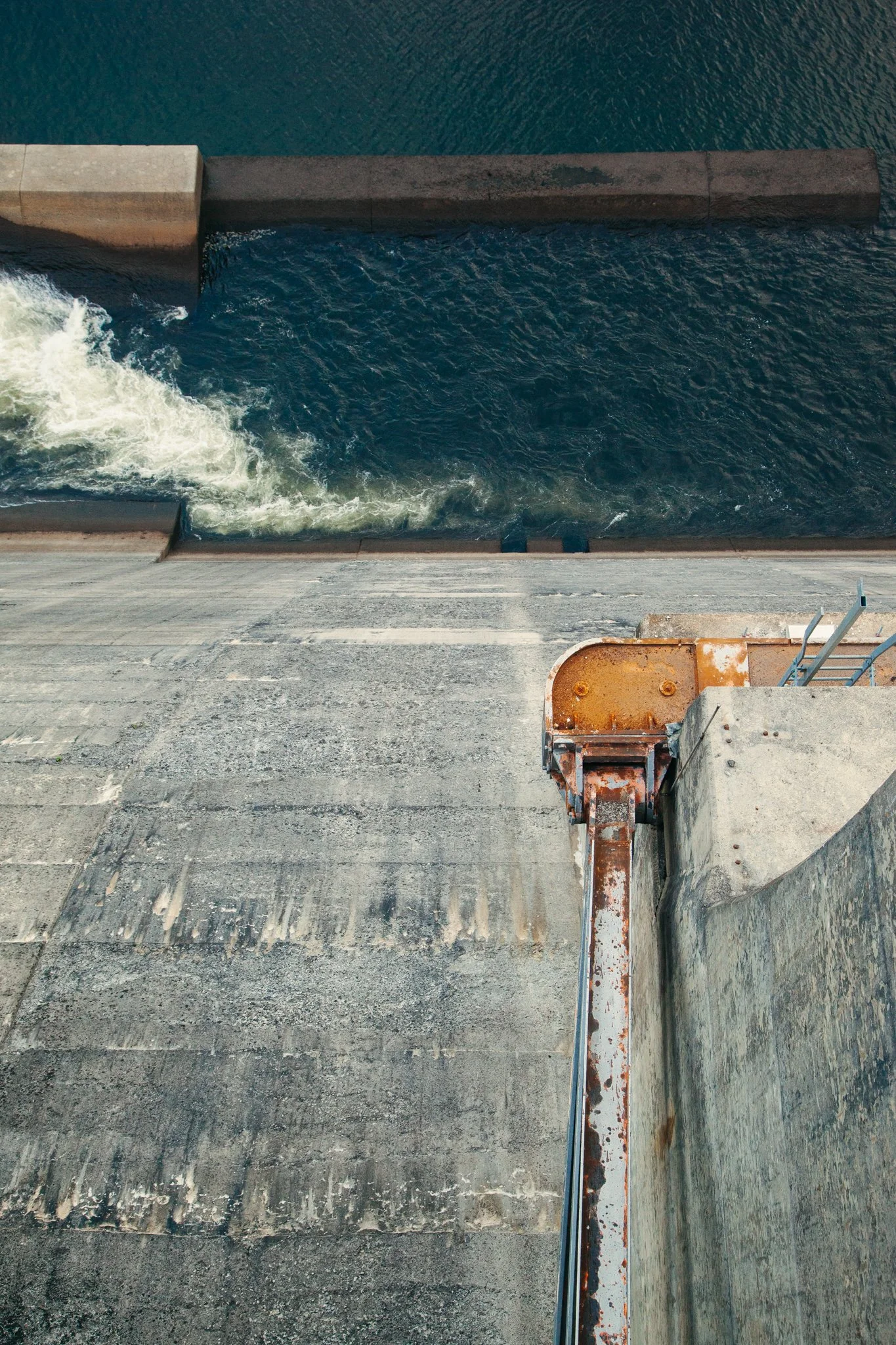 A top-down view of a dam spillway with water flowing through it, surrounded by concrete and a metallic railing, with an industrial area below.